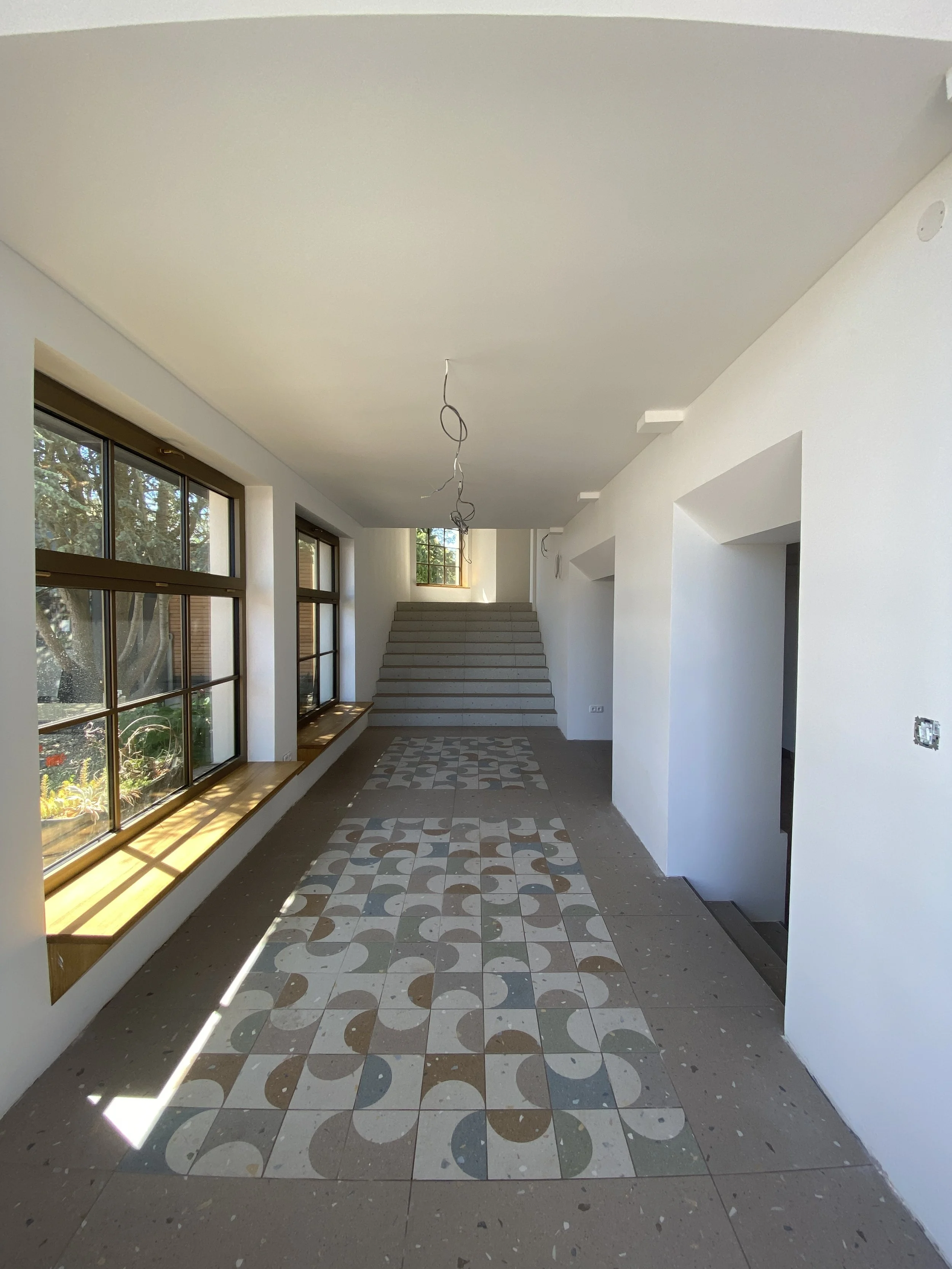 An indoor space with large windows on the left, white walls, a staircase at the back, and a patterned tile floor.