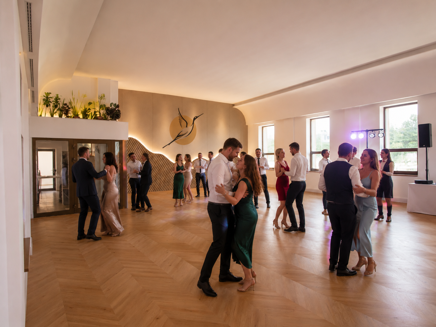 People dancing in pairs at a party or wedding reception in a brightly lit hall with wooden flooring and large windows.