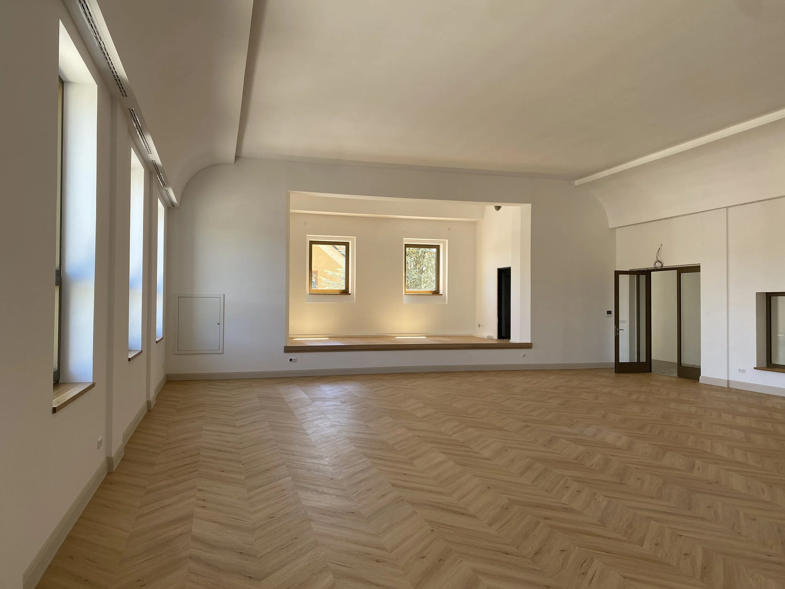 Empty spacious room with light-colored wooden floors, white walls, multiple windows, and a raised platform area.