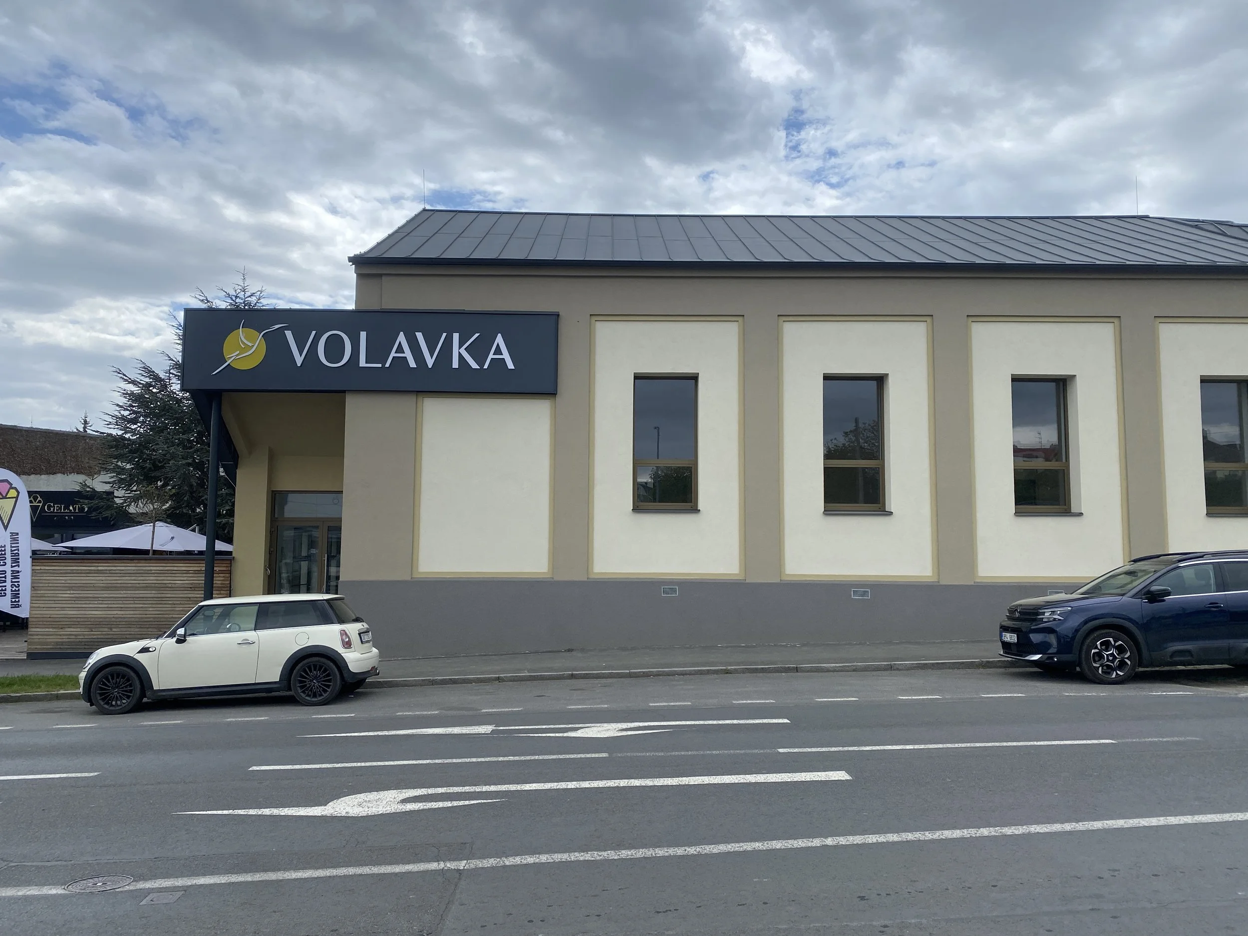 Photo of a building with a sign that says 'VOLAVKA', cars parked along the street in front, with a cloudy sky above.