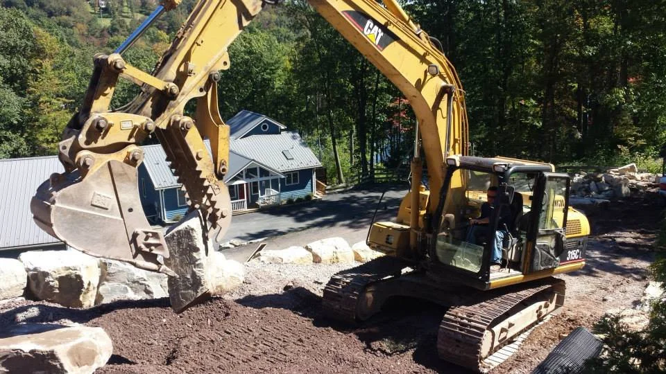 A yellow Caterpillar excavator moving dirt and large rocks on a construction site with trees and a house in the background.