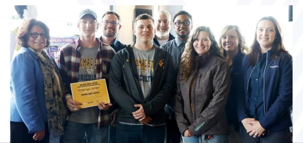 Group of people at a school event, some holding a yellow certificate, in an indoor setting with posters and windows in the background.