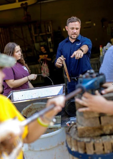 A man in a blue shirt cooking on a grill while children watch, at an outdoor event.