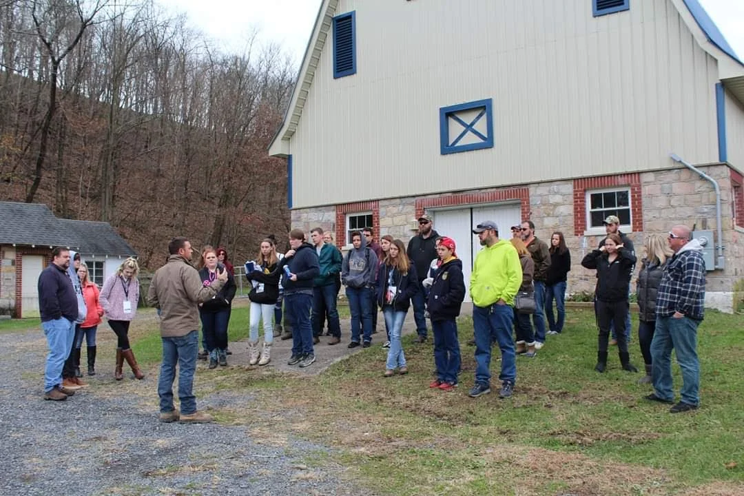 A group of people gathered outside a barn-like building with a wooded hill in the background, listening to a man in a brown jacket speaking.