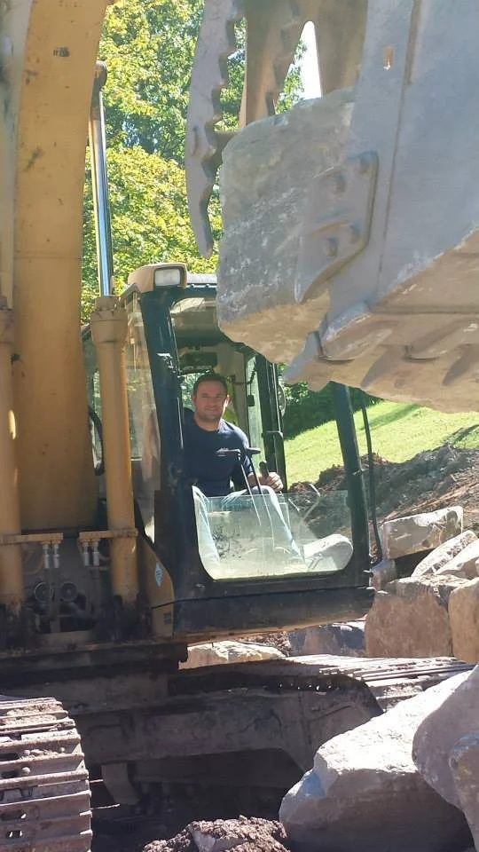 A construction worker operating a backhoe loader excavator amidst large rocks and boulders outdoors on a sunny day with greenery in the background.