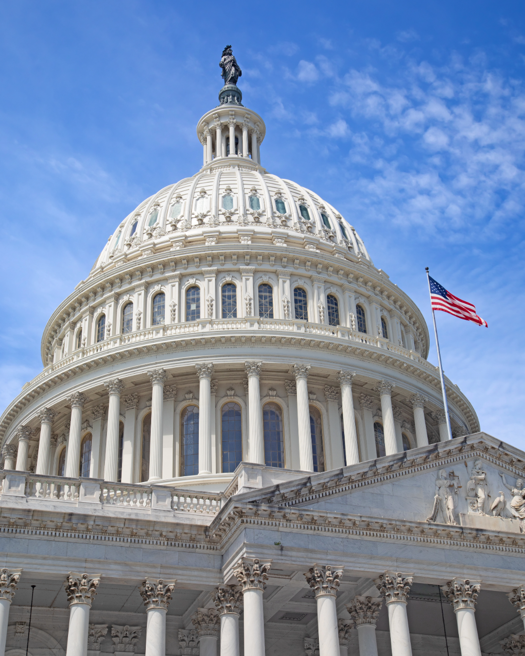 The United States Capitol building with its iconic dome, columns, and an American flag, under a blue sky with some clouds.