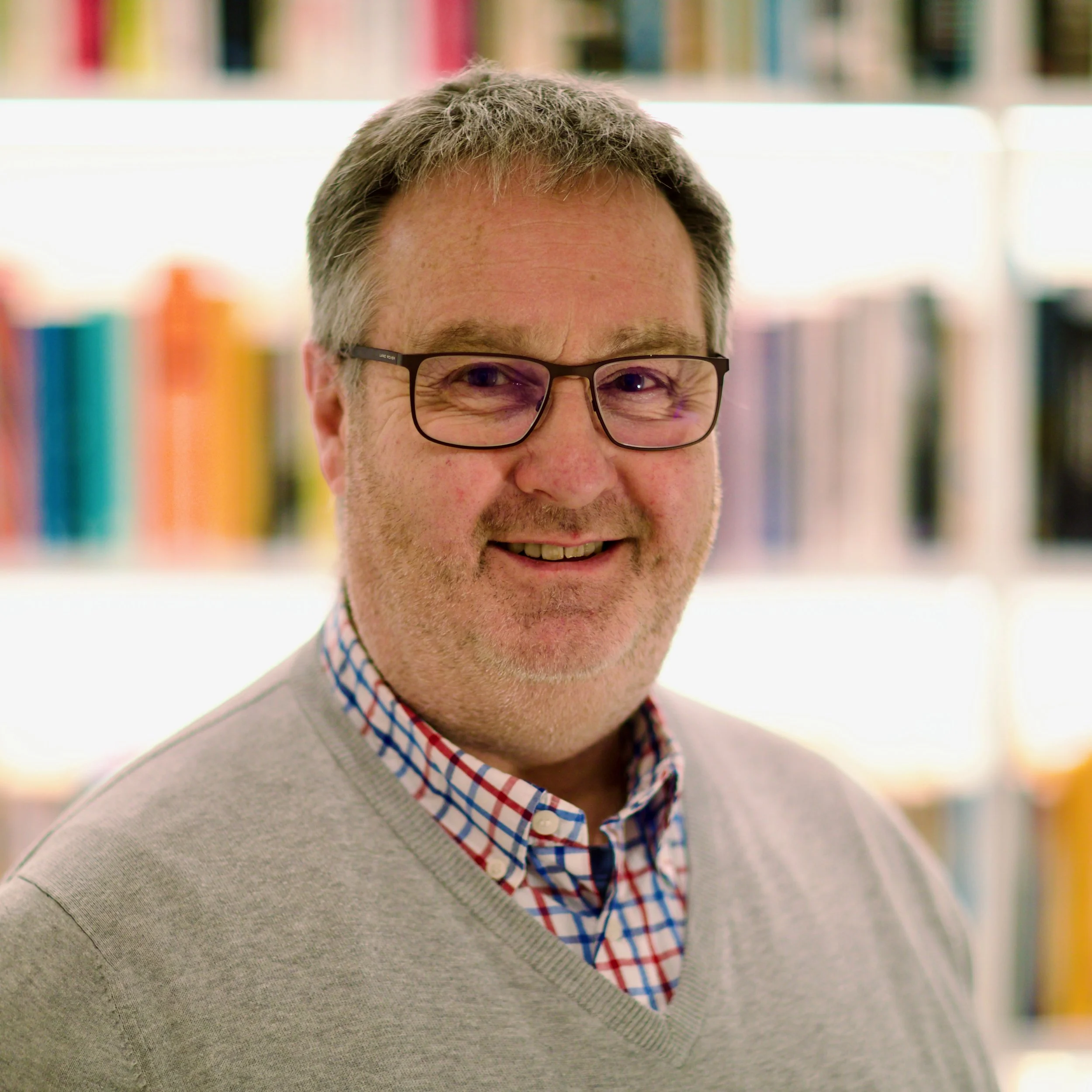 Smiling middle-aged man wearing glasses, a gray sweater, and a plaid shirt in a brightly colored office