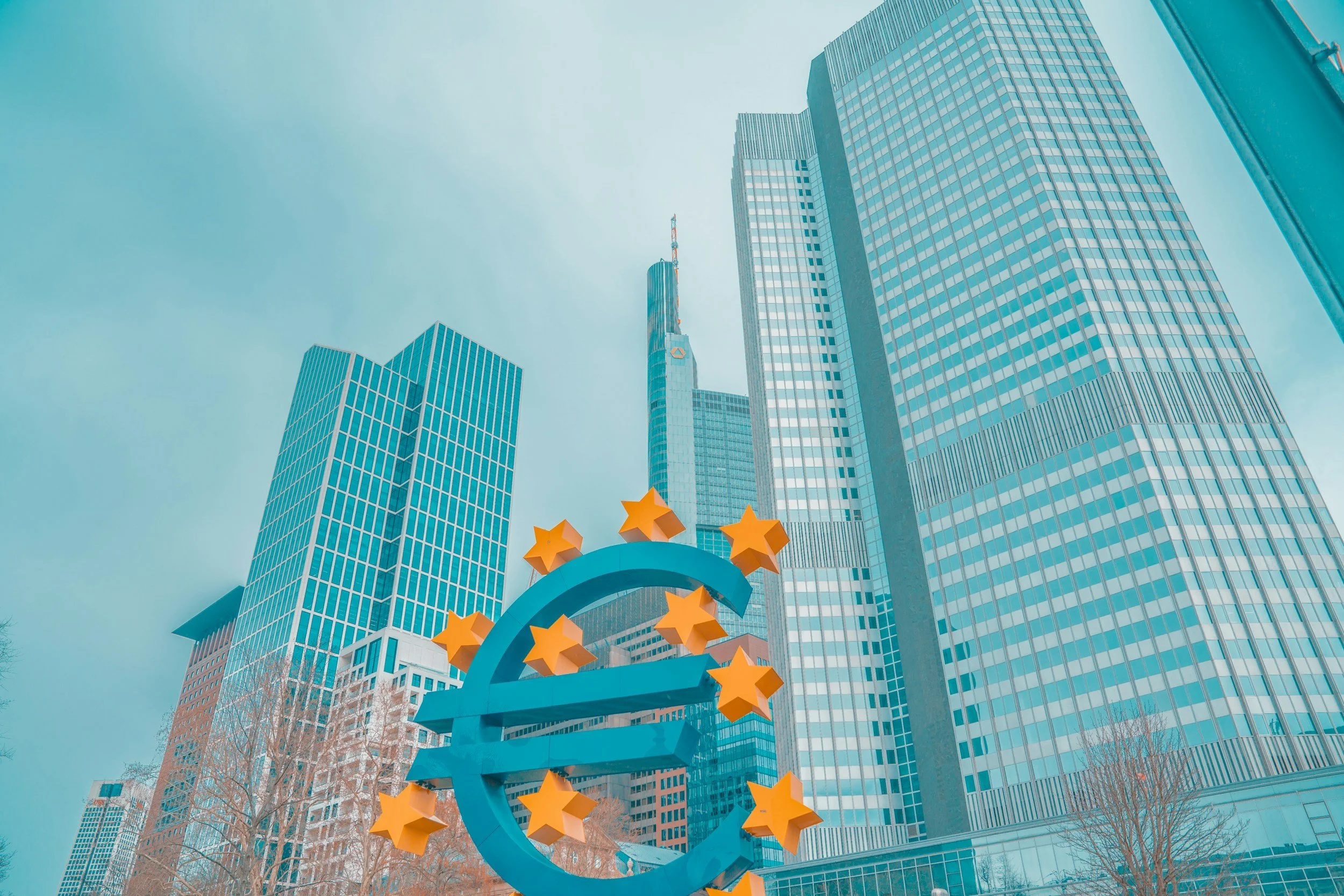 A large euro symbol sculpture with orange stars in front of modern skyscrapers in a cityscape.