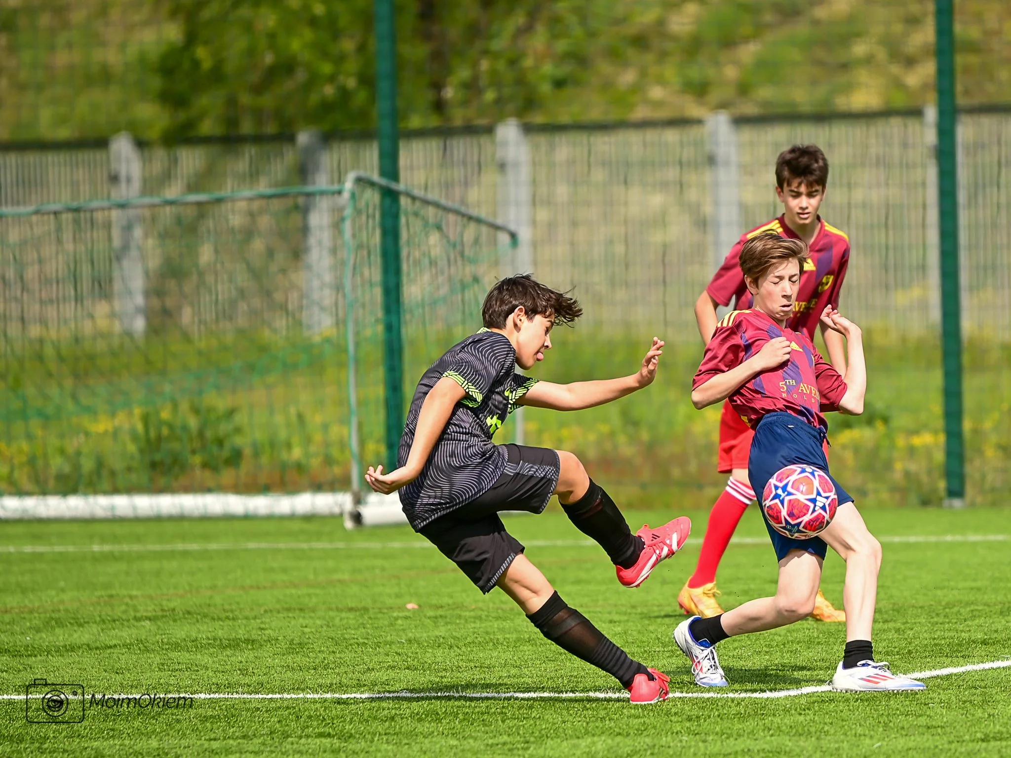Three boys playing soccer, two wearing red jerseys and one in black, on a green field with a fence in the background.