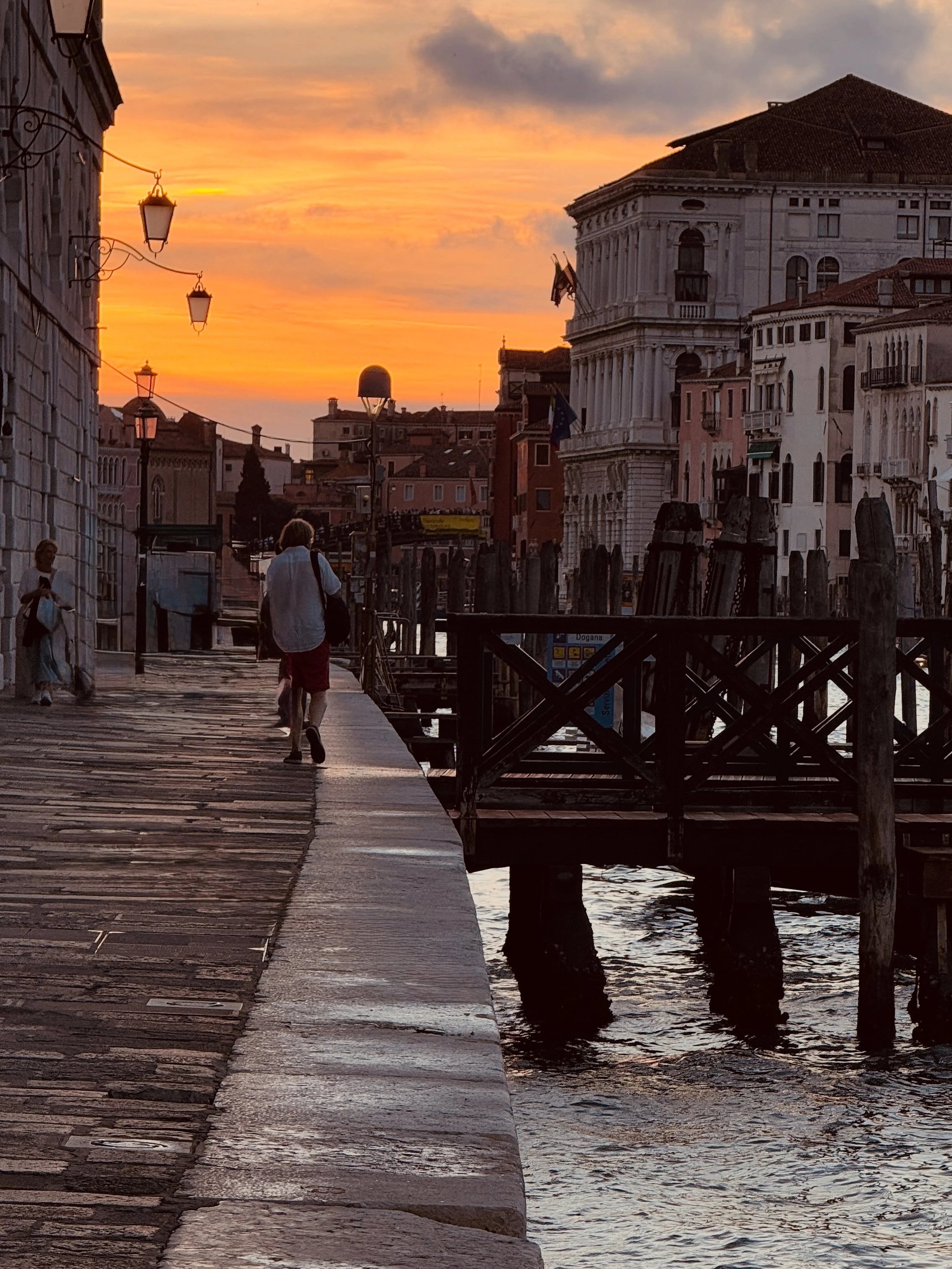 Venice, Italy waterfront walkway during sunset with historic buildings, lampposts, and a person walking along the canal.
