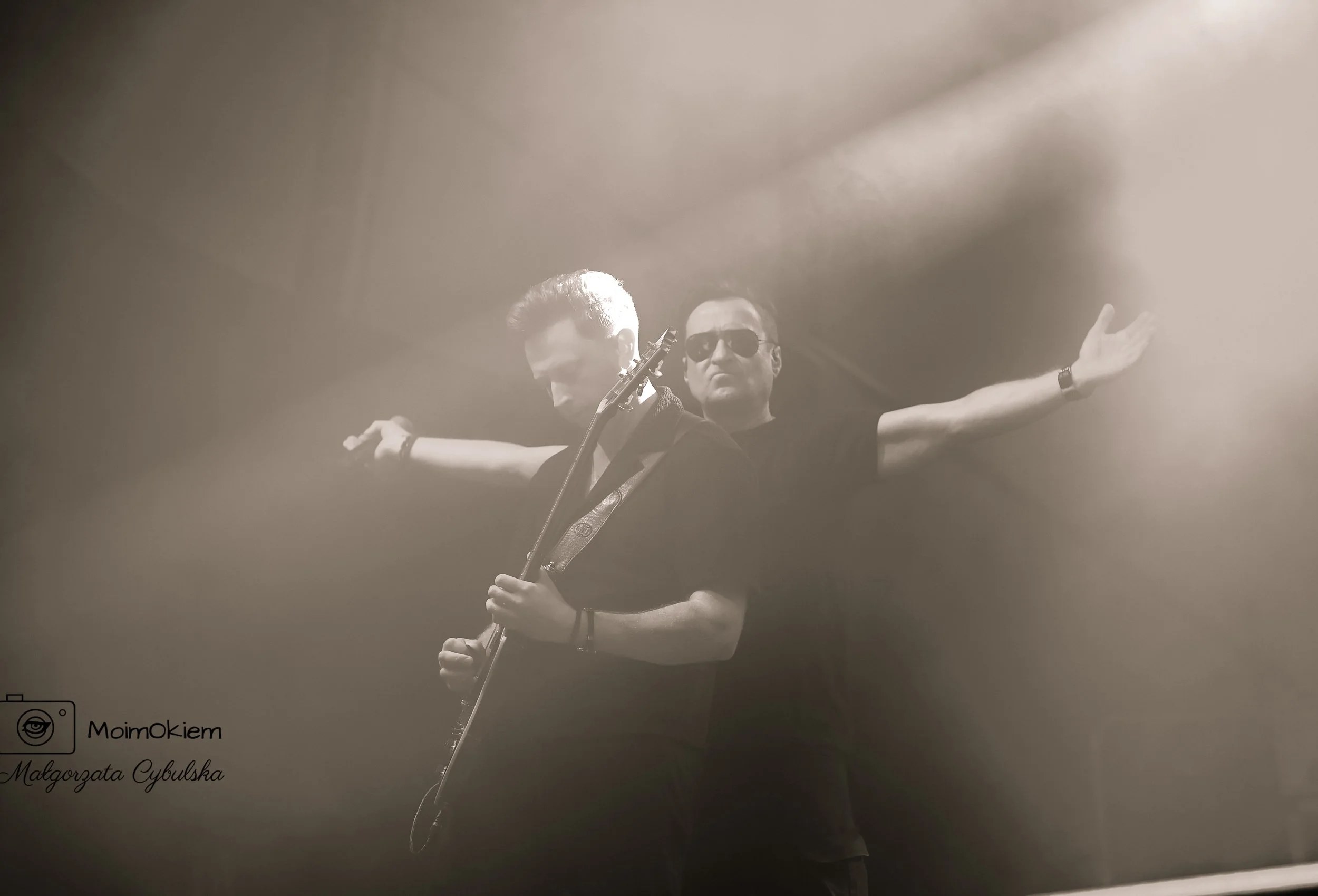 Two men performing on stage under dramatic lighting, one playing an electric guitar with arms extended, the other standing behind with sunglasses and arms spread wide.