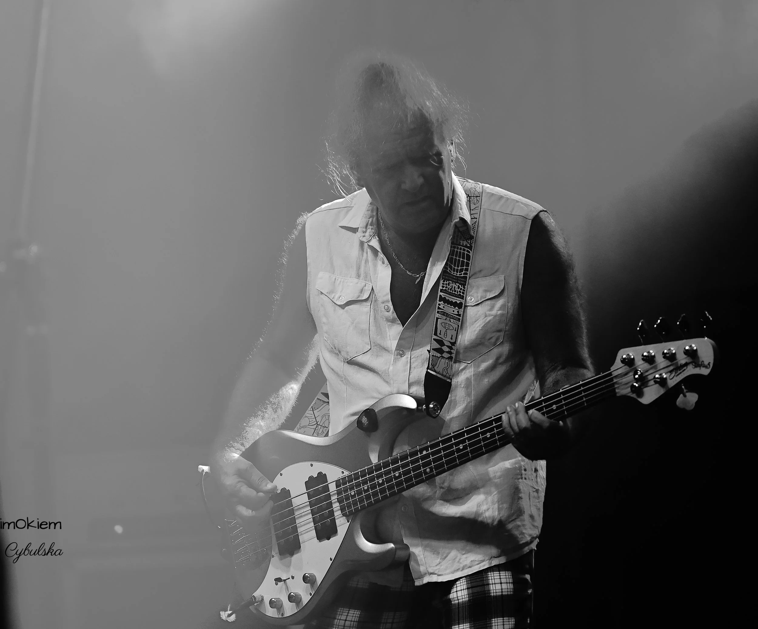 A black-and-white photo of a man with messy hair playing an electric bass guitar on stage. He is wearing a sleeveless shirt and plaid shorts, with a patterned strap on the guitar. The stage background is dark, and light is shining from above.