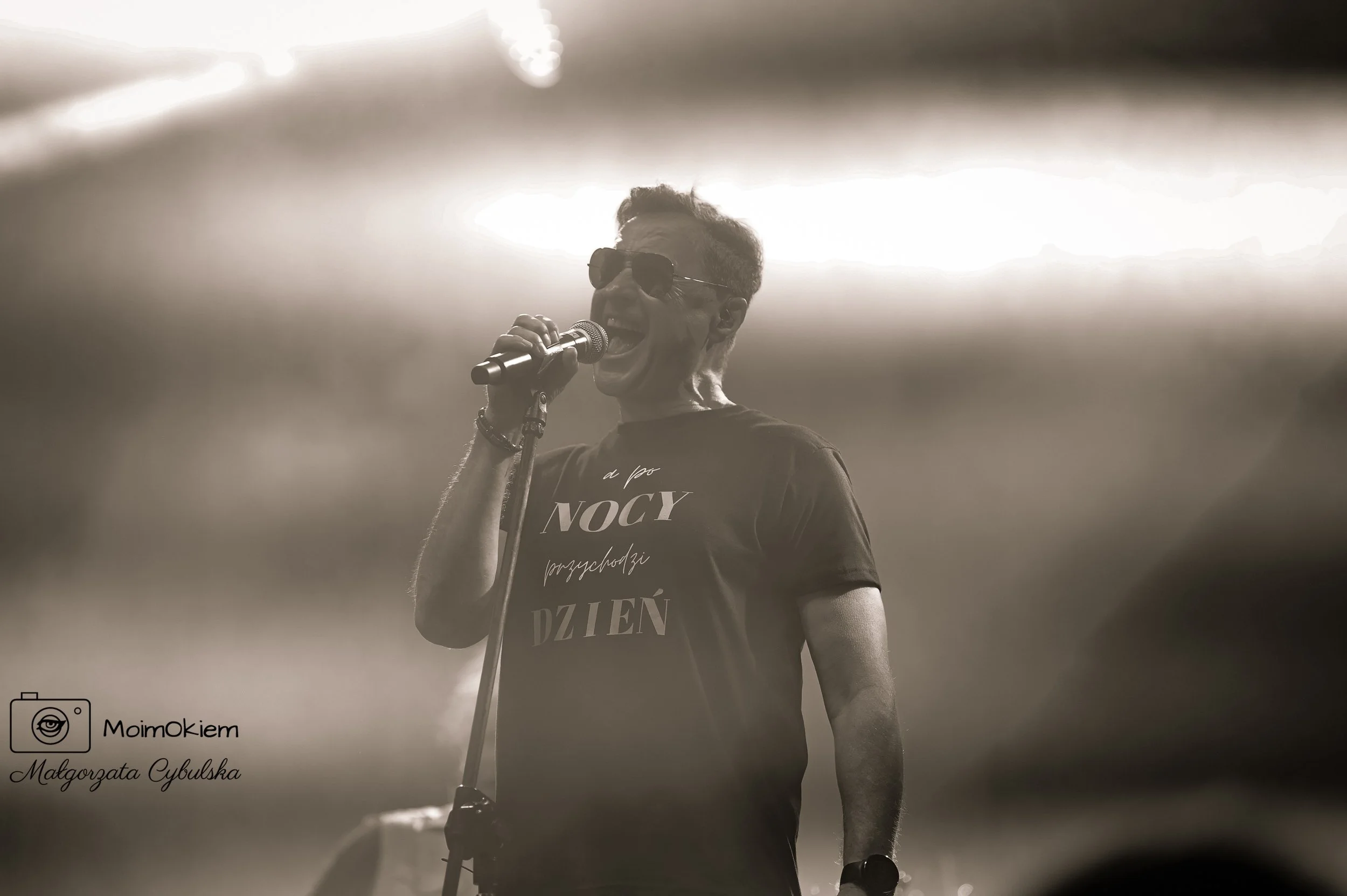 A man singing into a microphone on stage, wearing sunglasses and a T-shirt with Polish text, during a performance with bright lights in the background.