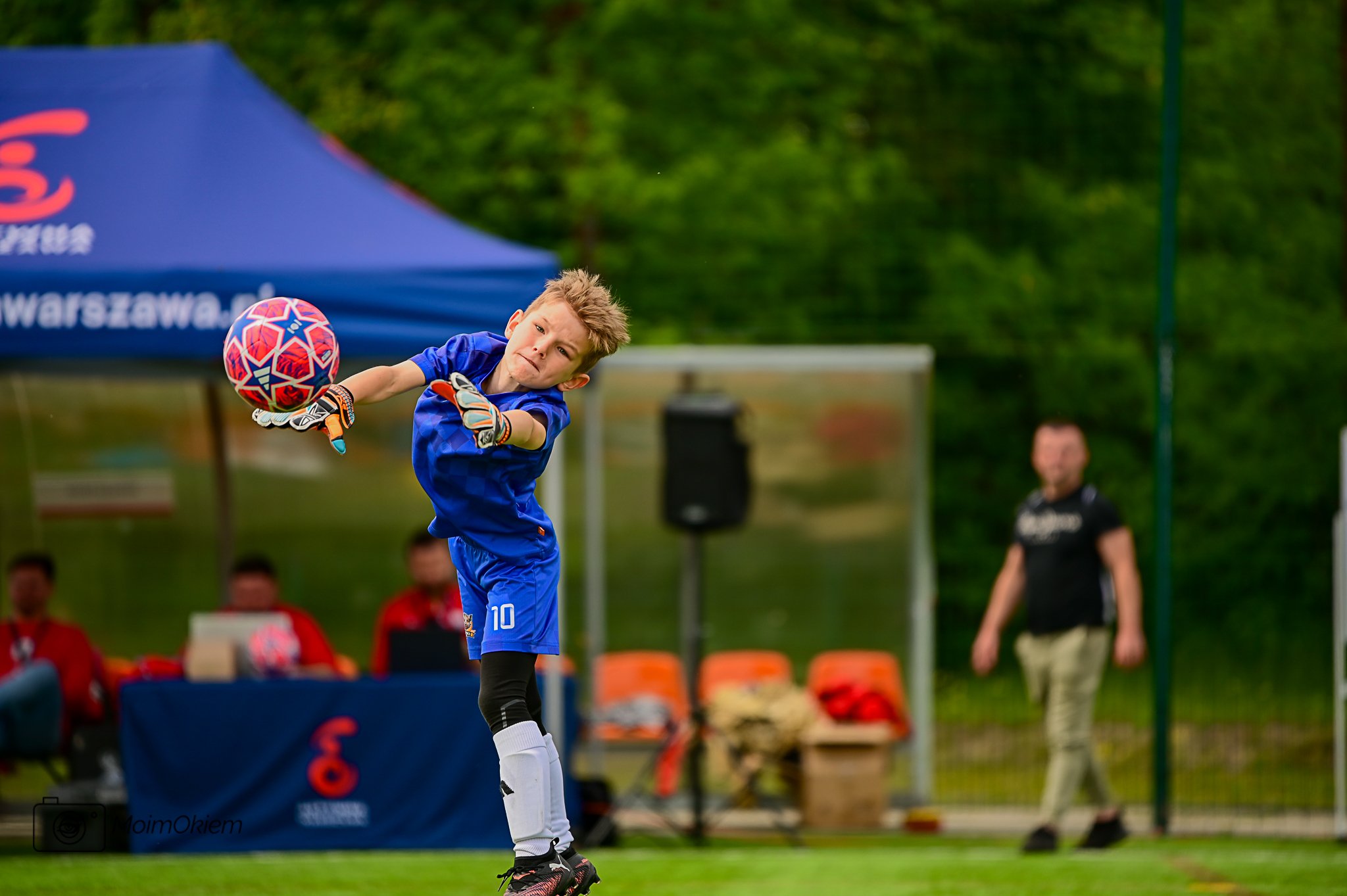 A young boy in a blue soccer uniform and gloves is jumping to catch a colorful soccer ball during a game on a grassy field, with a blue tent and other people in the background.