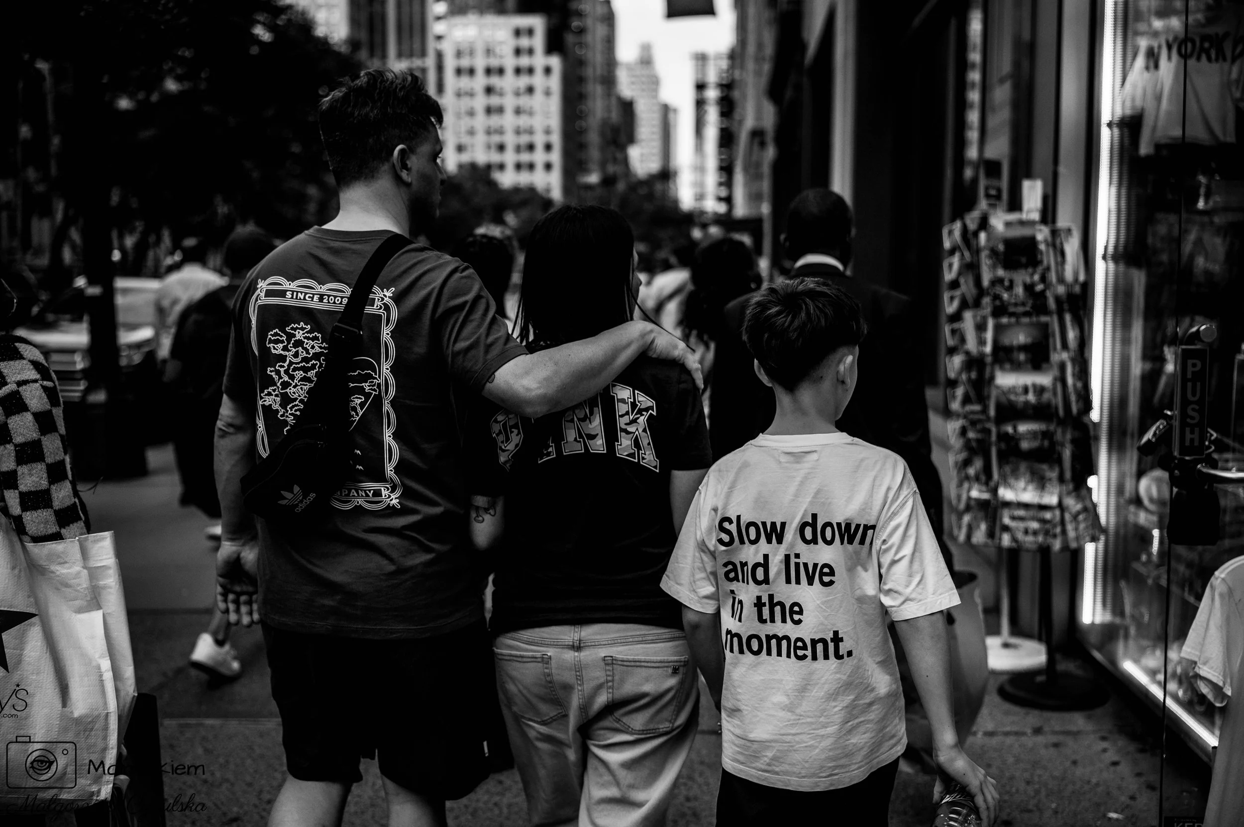 People walking on a busy city sidewalk, with tall buildings in the background. A man has his hand on a woman's shoulder, and a young boy nearby wears a t-shirt that says 'Slow down and live in the moment.'