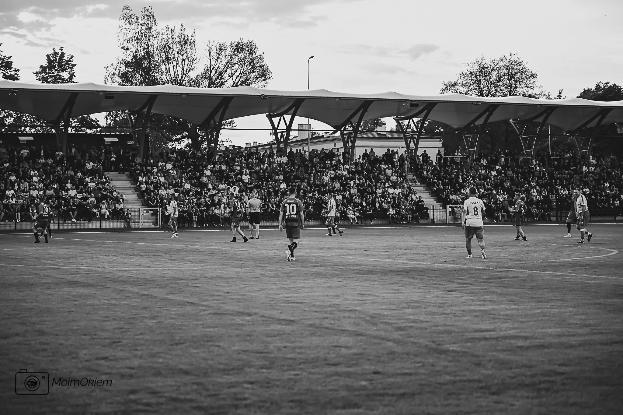 Black and white photo of a soccer match at a stadium with people in the stands and trees in the background.