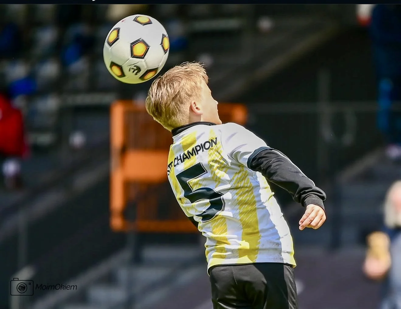 Young soccer player with blonde hair in a yellow and white jersey jumping to head the soccer ball during a game.