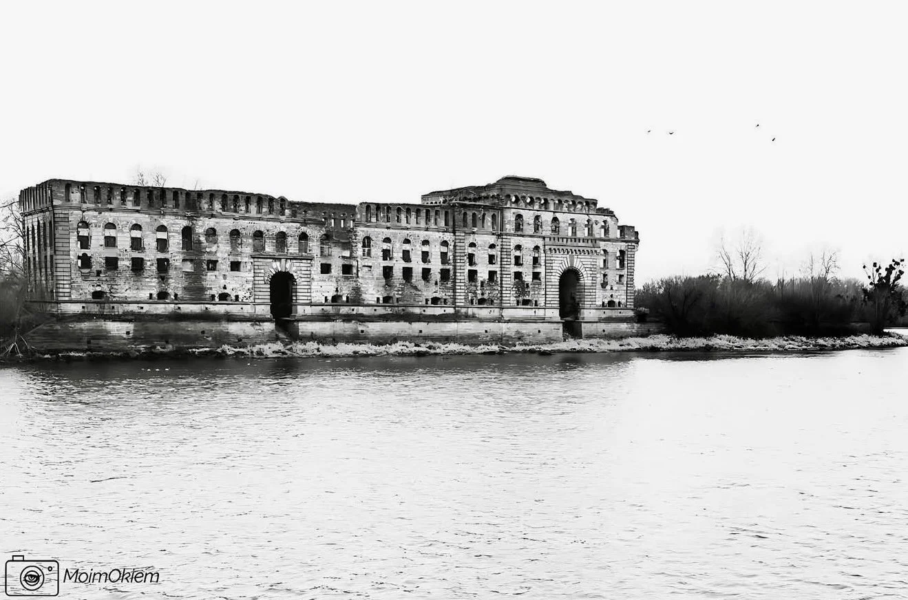 Black and white photo of an old, abandoned fortress or castle on the riverbank, with visible deterioration and broken windows, surrounded by leafless trees.