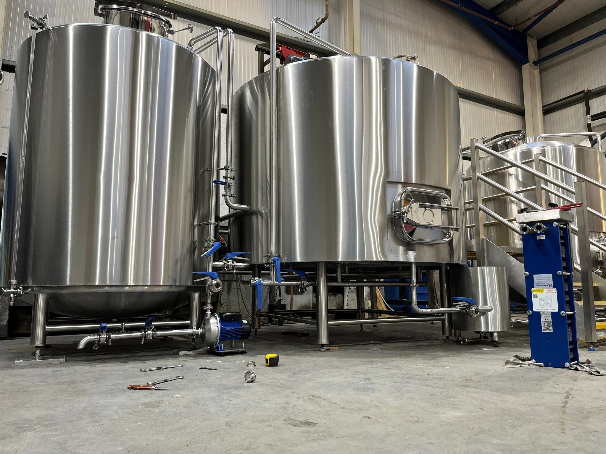 Large stainless steel industrial tanks and equipment in a manufacturing facility, with tools and pipes on the floor.