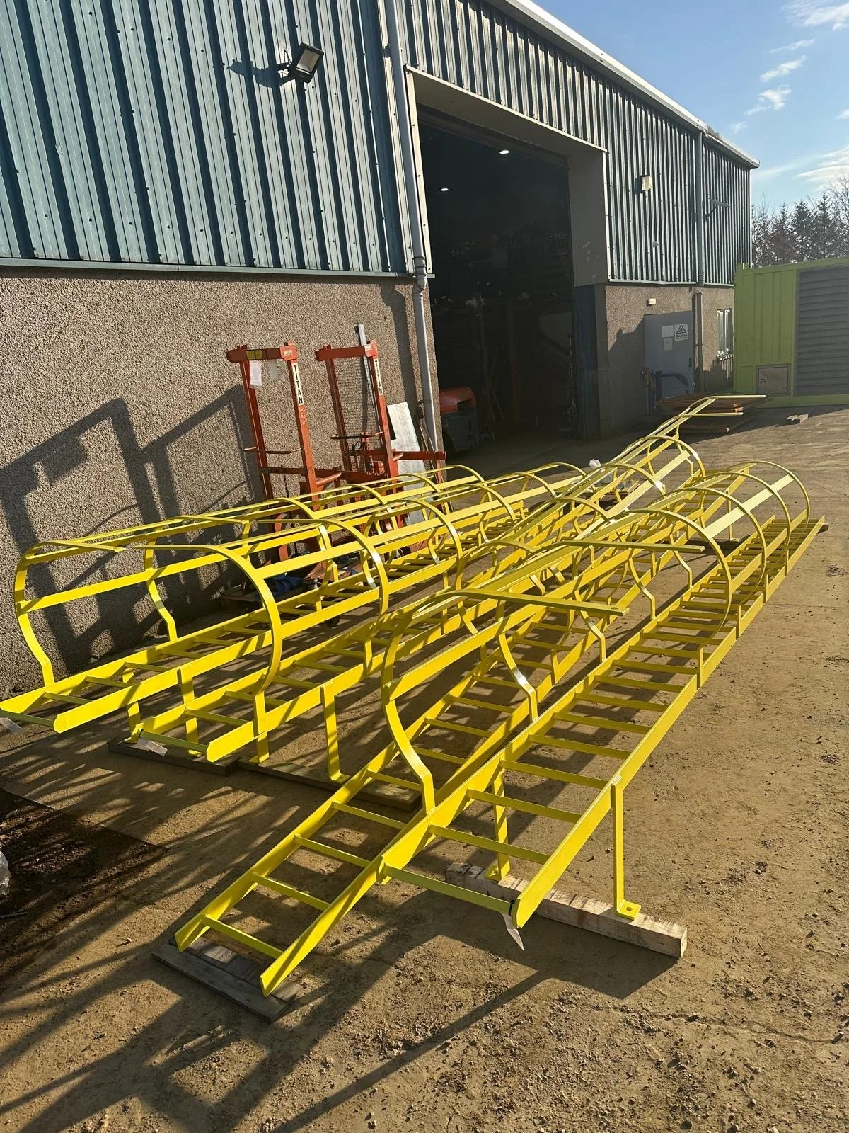 Yellow outdoor staircase frames with curved tunnels, placed on wooden planks outside a building with metal siding and a large open garage door.