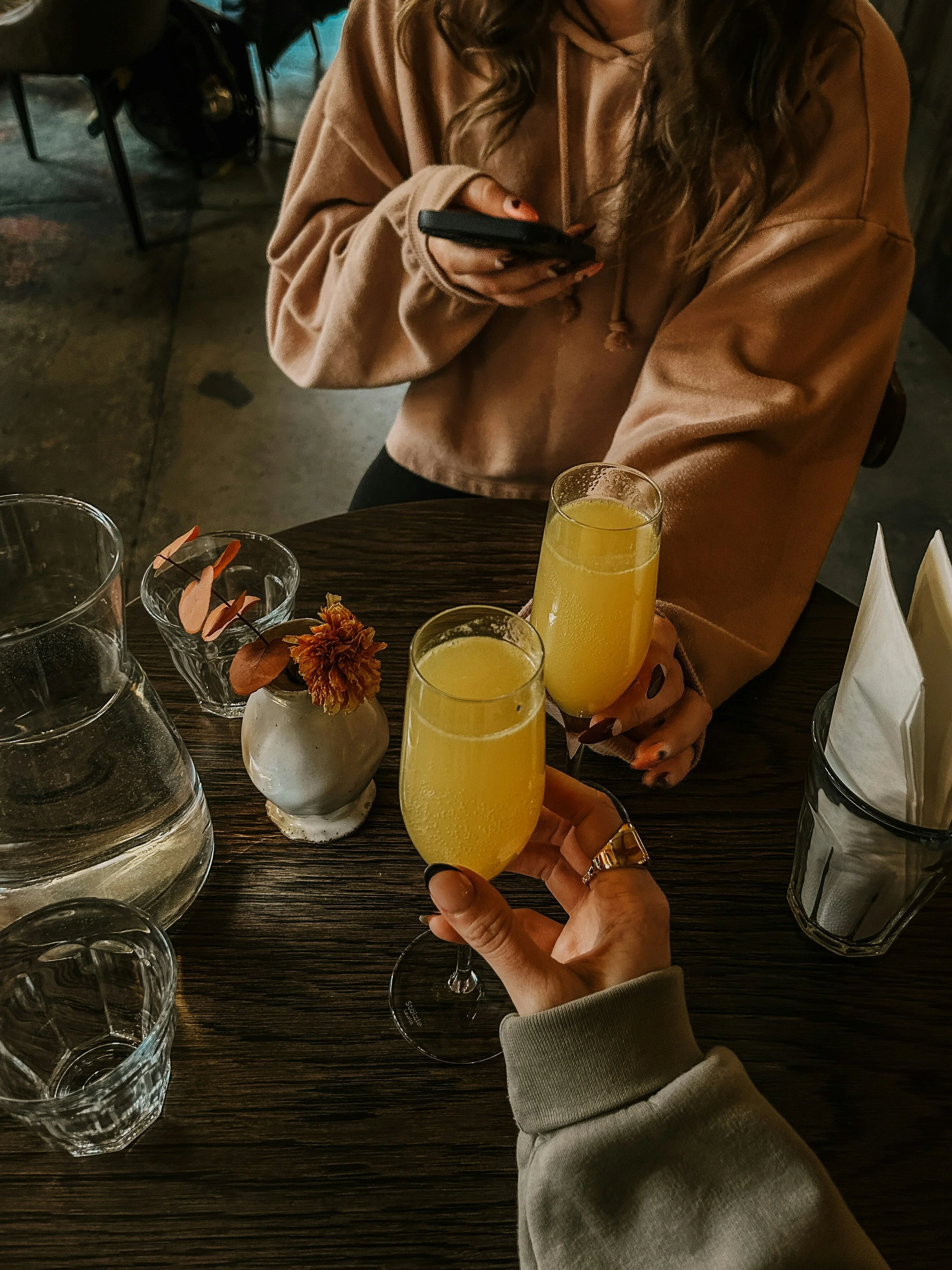 Två personer som sitter vid ett bord med frukostdrycker i champagneglas, tårta i vas och vattenkaraff, en person tar en selfie och den andra håller ett glas.