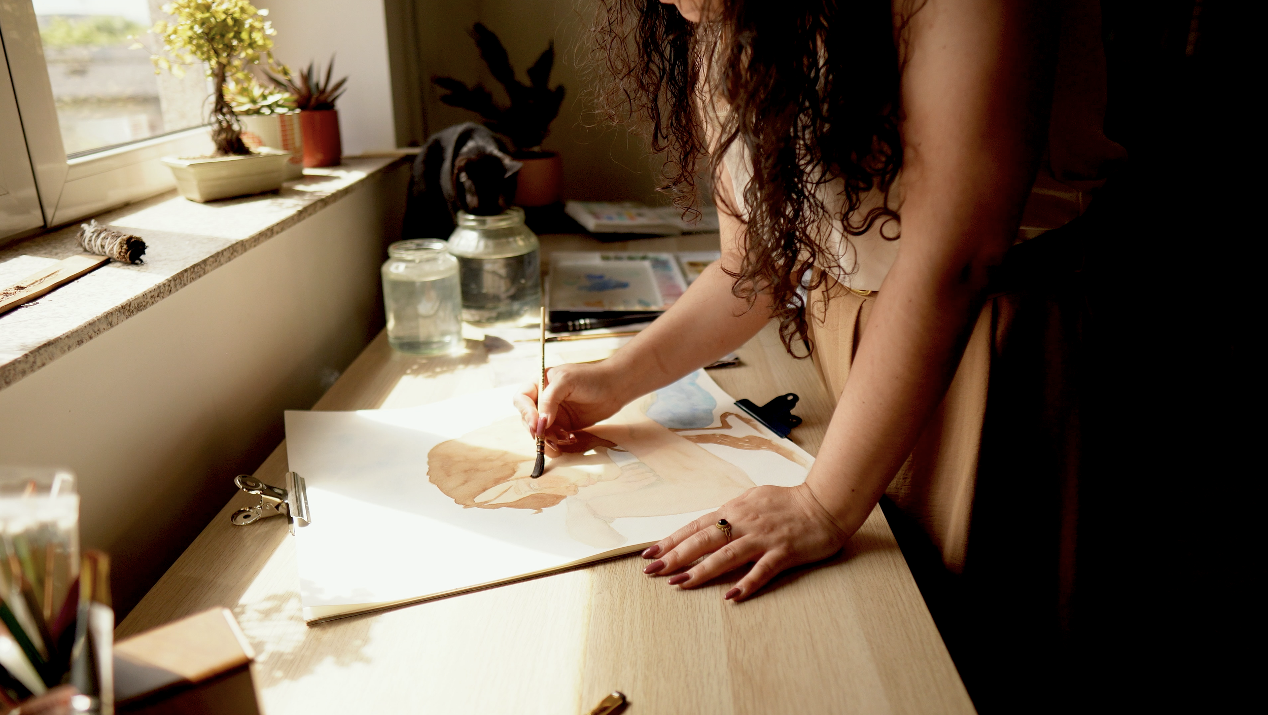 A person with curly hair painting a watercolor picture on a large sheet of paper at a wooden desk in a sunlit room, with a black and white cat on the windowsill observing.
