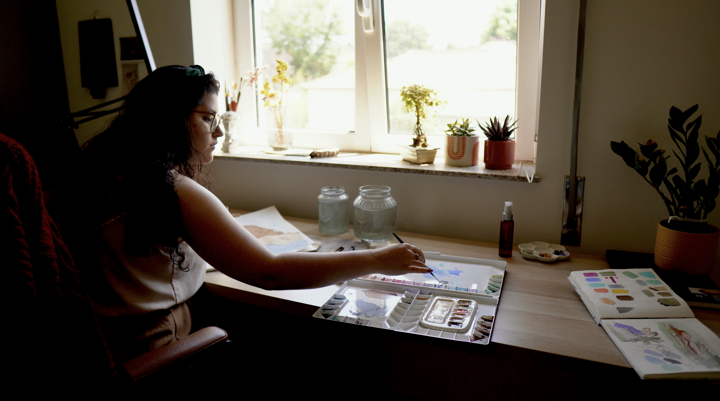 A woman sitting at a desk painting with watercolors, surrounded by art supplies and potted plants near a window.