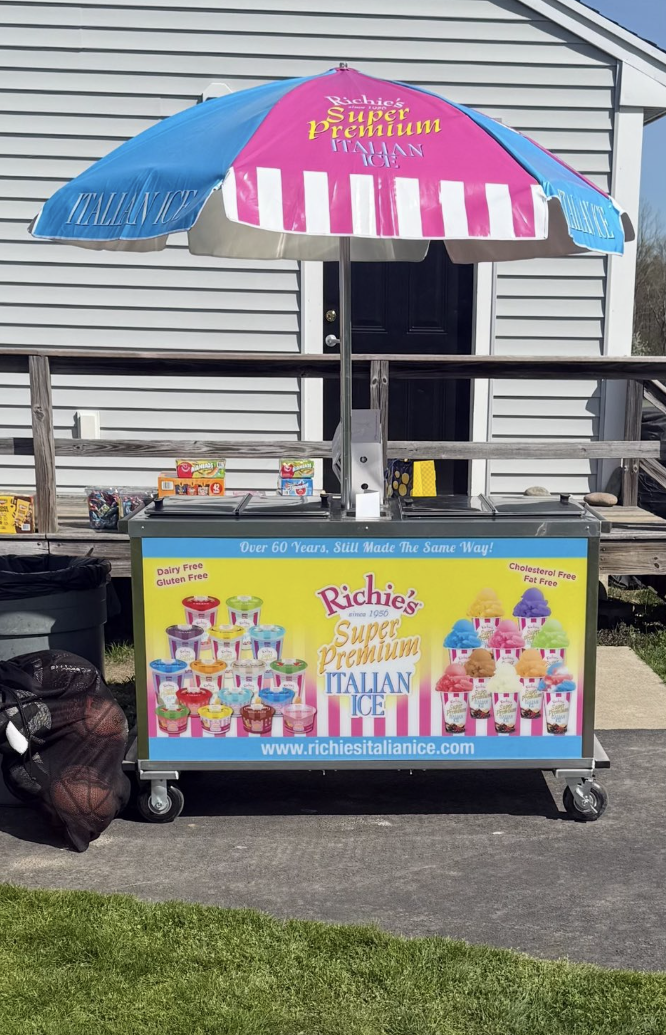 An ice cream cart advertising Richie’s Super Premium Italian Ice, featuring a large pink and blue umbrella and a colorful poster with various ice cream flavors and website URL.
