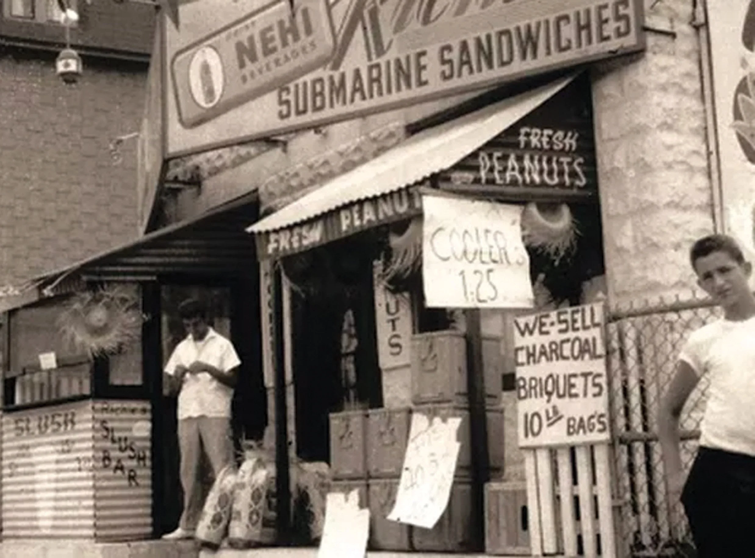 A black-and-white photo of a street scene with a storefront selling food items like peanuts, roasted chestnuts, and charcoals. There are several hand-painted signs with prices and product names hanging on or near the storefront, and a man is standing in front of it.