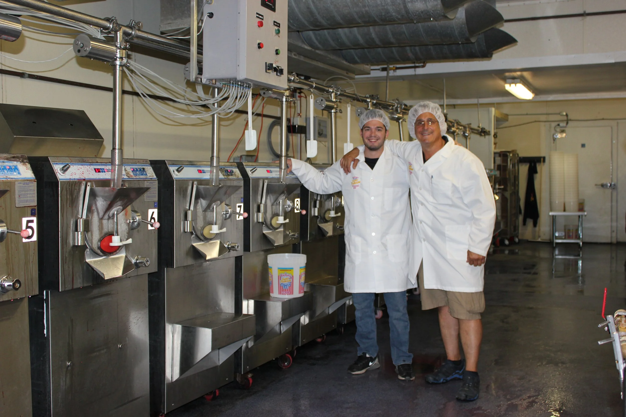 Two men standing next to a row of cotton candy machines in a factory or food production setting, with one man in a white coat and hairnet, smiling.