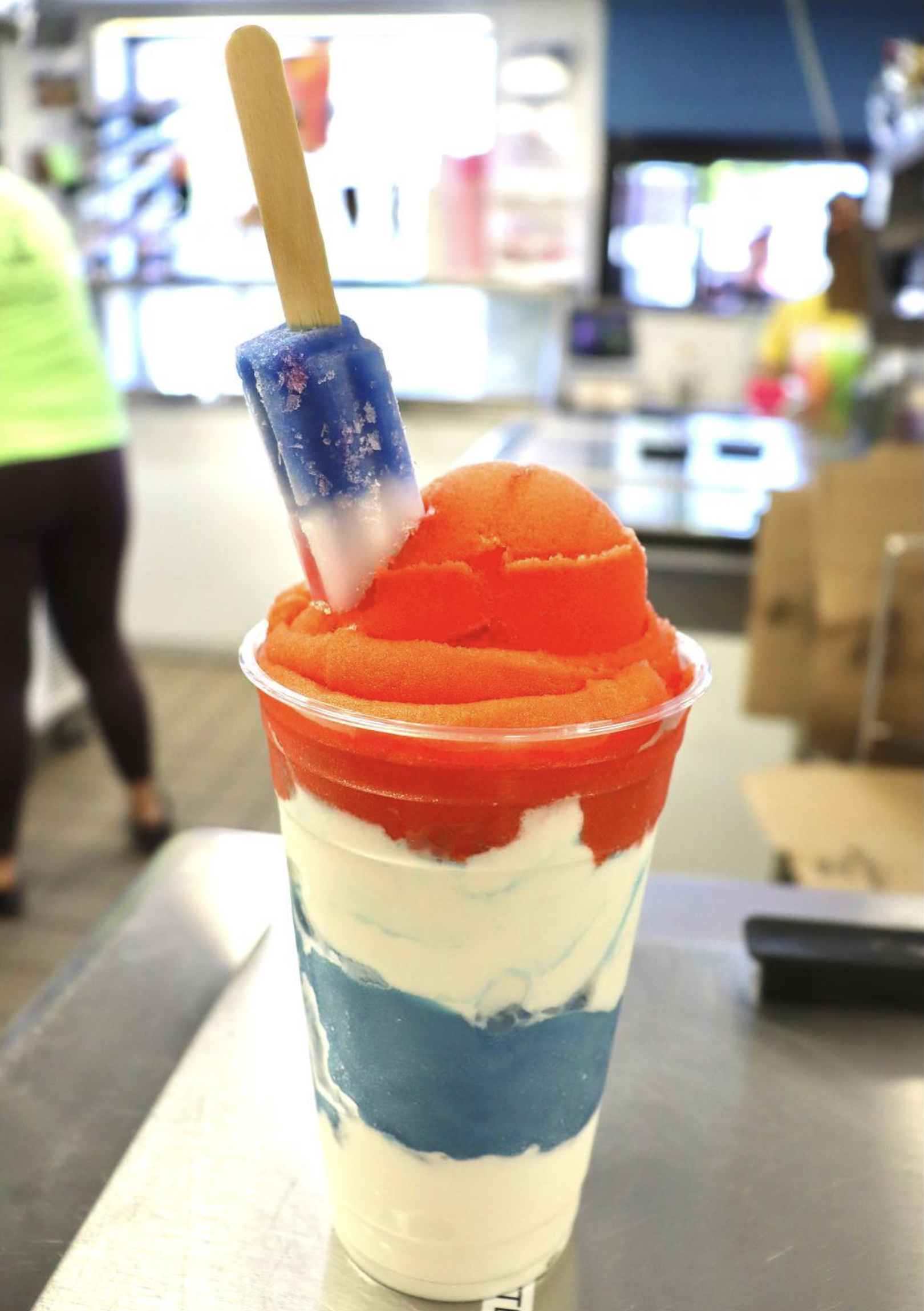 A cup of ice cream with orange and white swirls, topped with an orange scoop of sherbet and a blue popsicle, set against a blurred background of a shop or market interior.