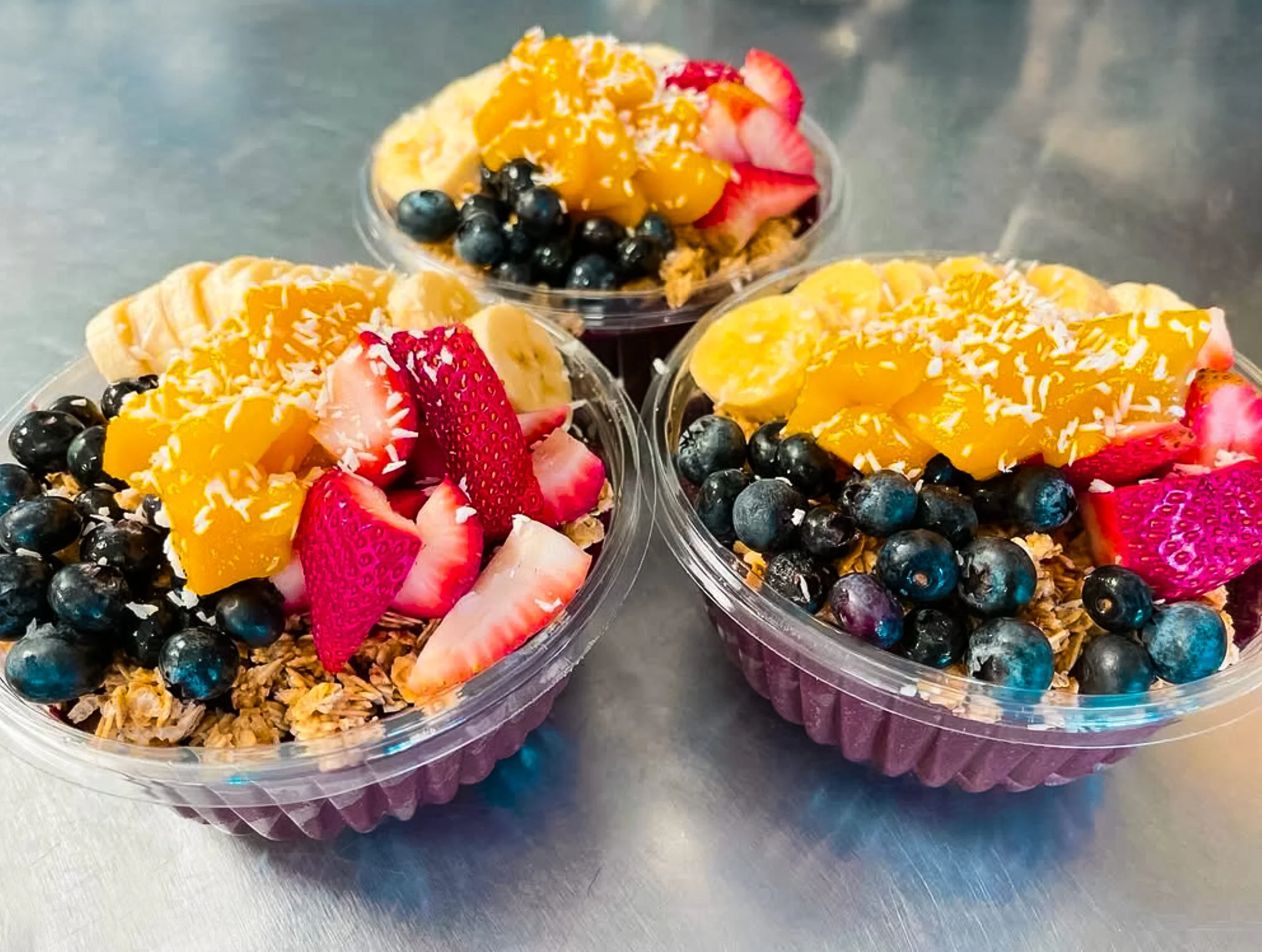 Two clear plastic bowls filled with granola, topped with sliced strawberries, blueberries, banana slices, and mango chunks.
