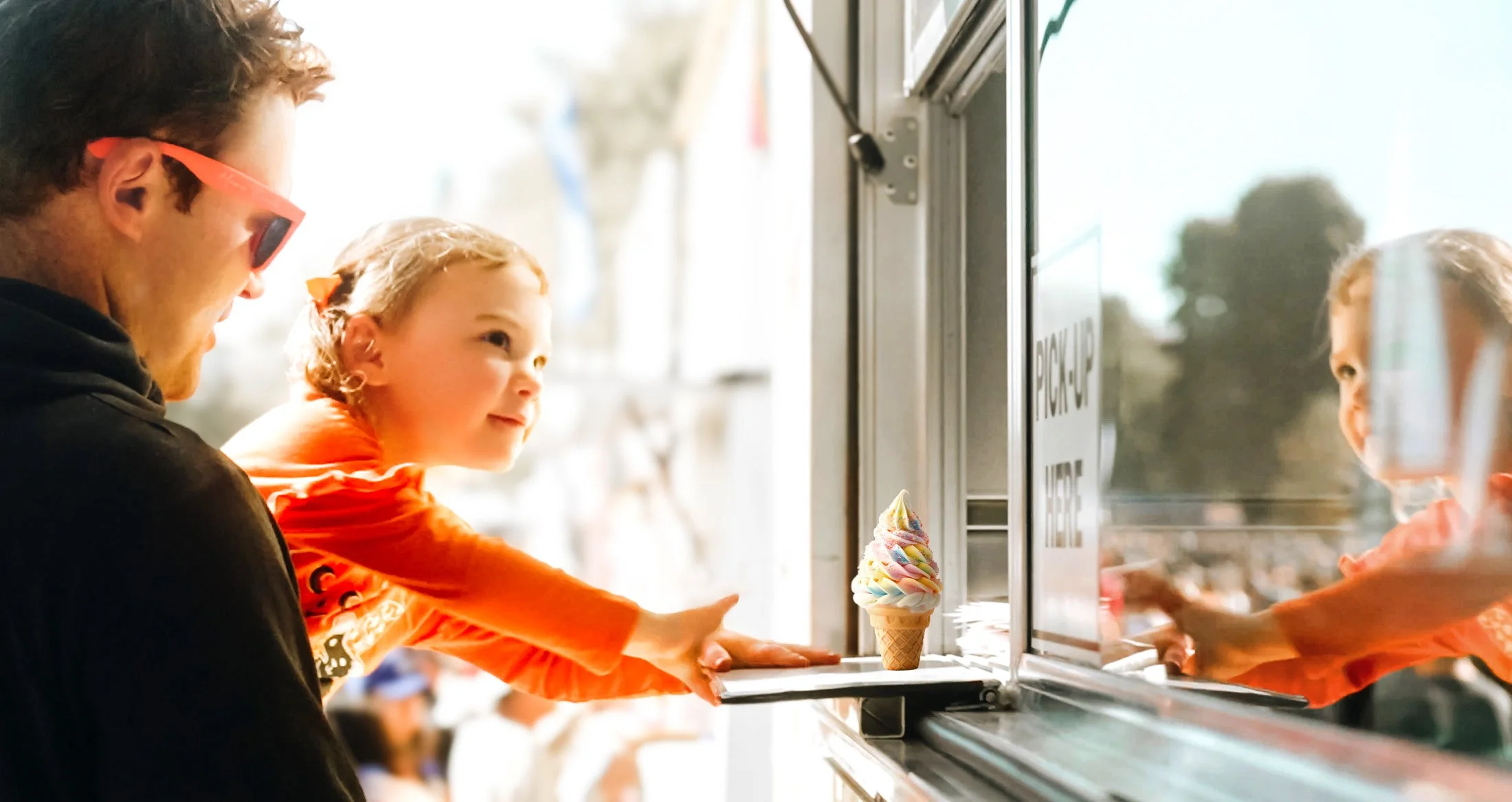 A man and young girl at an ice cream shop, reaching out for ice cream cones, with a sign on the counter and a phone booth in the background.