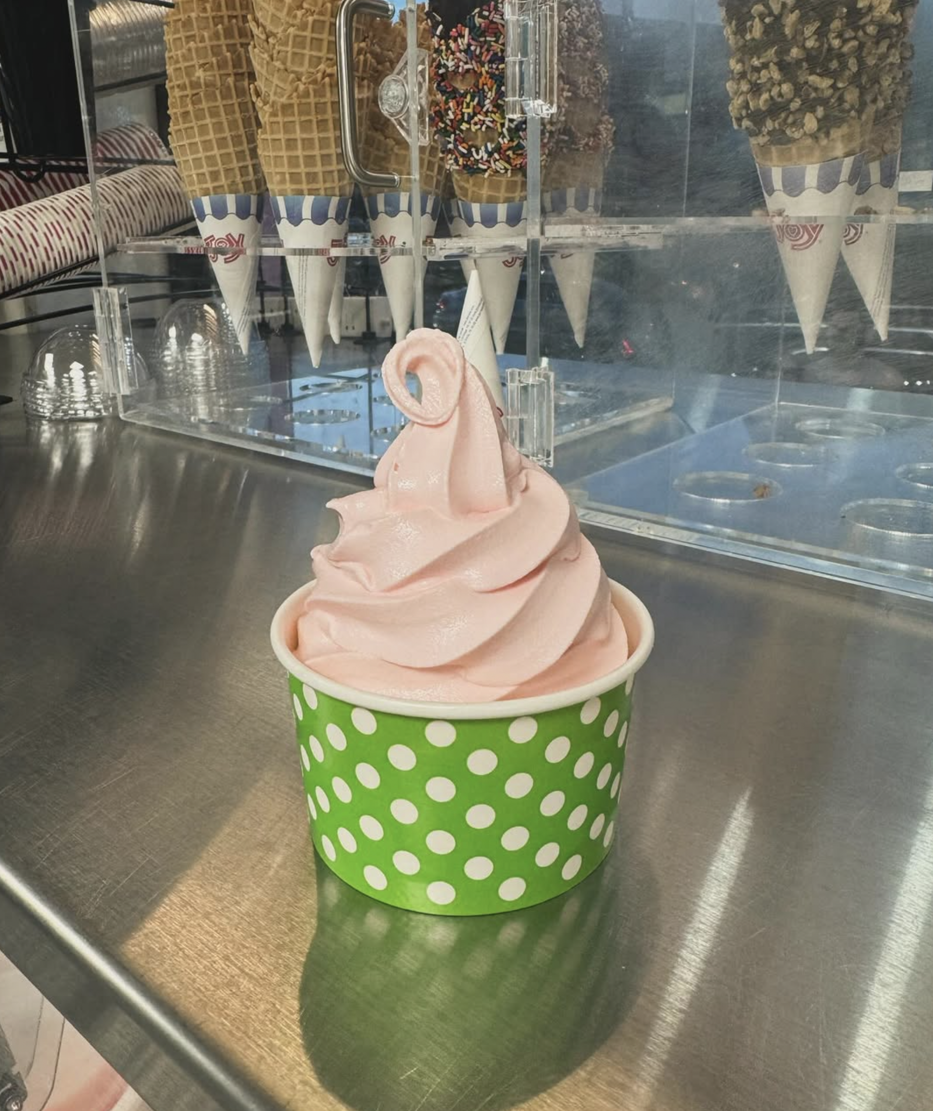 Ice cream cone-shaped container with pink soft serve ice cream topped with more ice cream, placed on a stainless steel surface in front of a glass display case filled with waffle cones, toppings, and sprinkles.