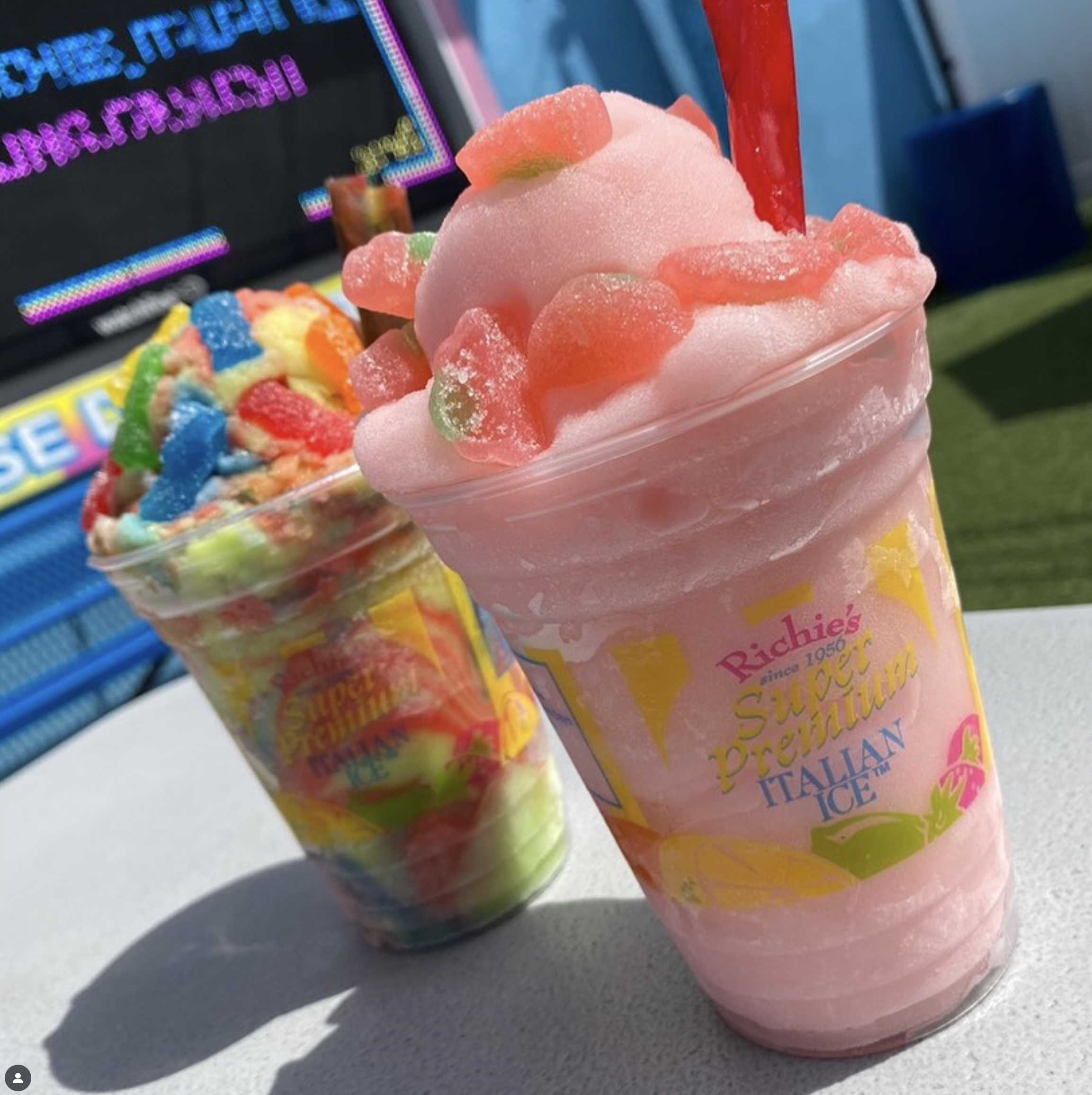 Close-up of a pink cotton candy in a transparent plastic cup with colorful gummy candies on top, with a red straw inserted. In the background, part of another colorful cotton candy and a sign with neon colors. The item is placed on a flat surface outdoors.