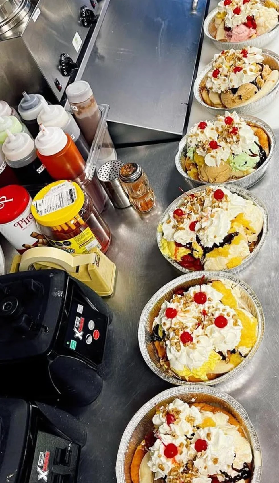 Multiple pies topped with whipped cream, cherries, caramel sauce, and nuts on a stainless steel counter in a dessert shop.
