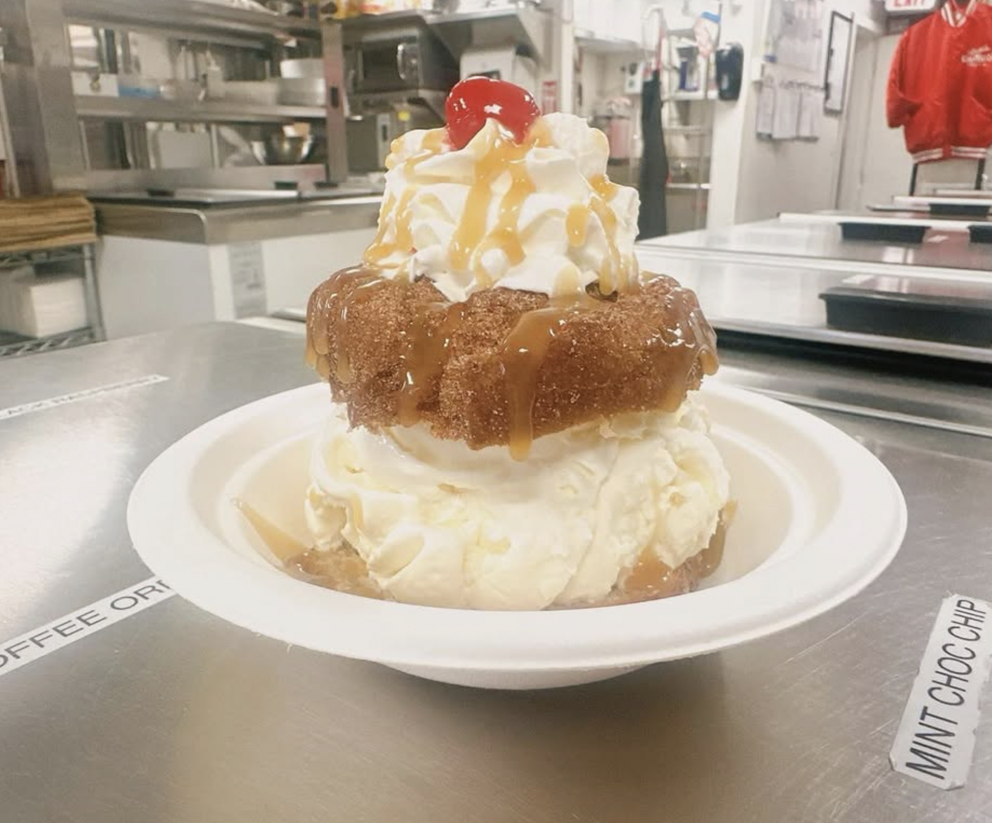 A dessert with a piece of fried chicken on top, whipped cream, caramel sauce, and a cherry garnished on the whipped cream, served on a white plate in a commercial kitchen or restaurant setting.