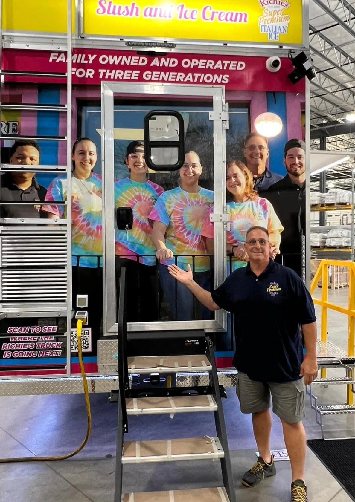 Photo of a mobile ice cream truck with nine employees, some of whom are wearing colorful tie-dye shirts. A man in shorts and a polo shirt is standing in front, smiling and gesturing towards the truck. The truck has a sign that reads "Family Owned and