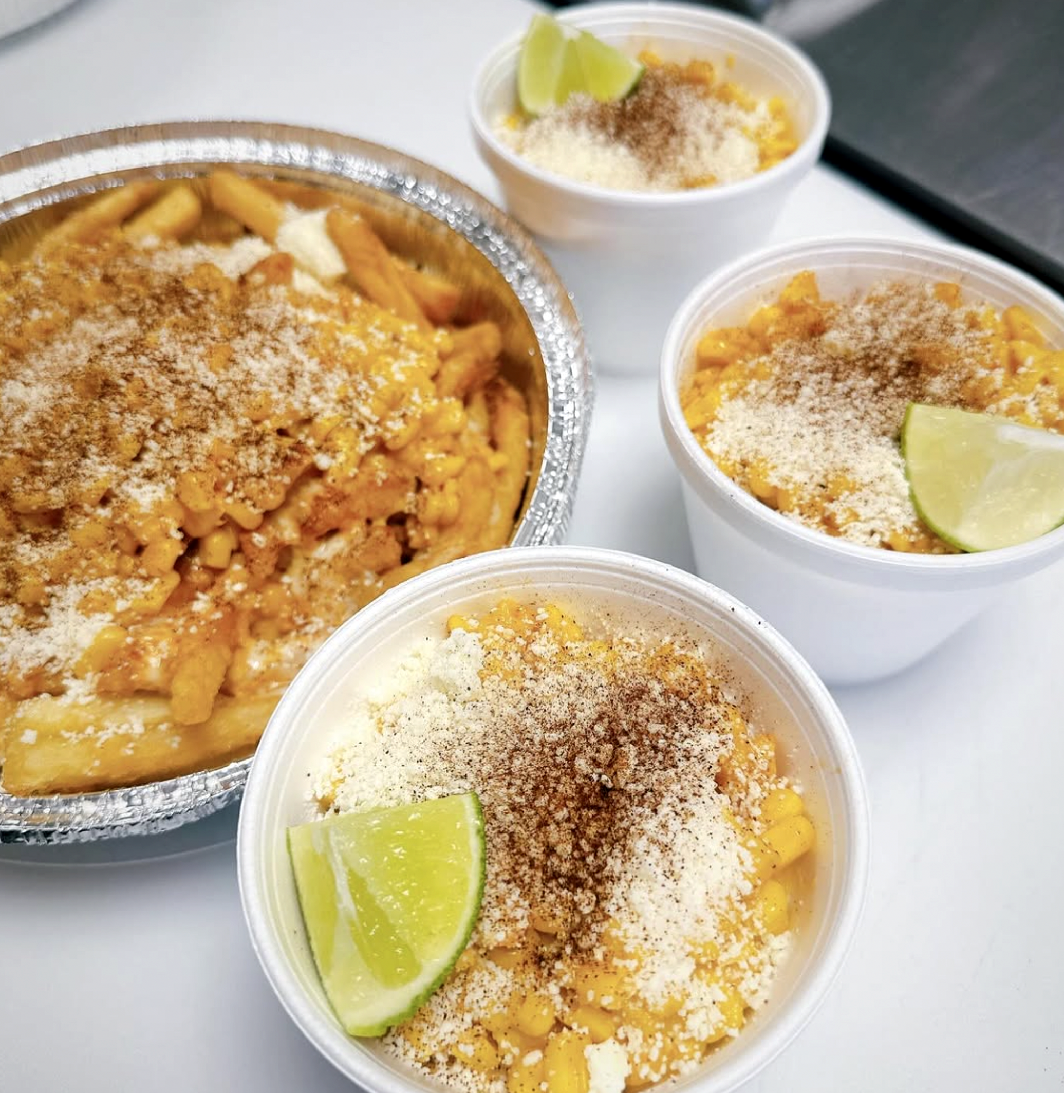 Close-up image of two plastic containers filled with Mexican street corn sprinkled with cheese and chili powder, along with one container of Mexican rice with cheese and lime wedges on top, on a white surface.