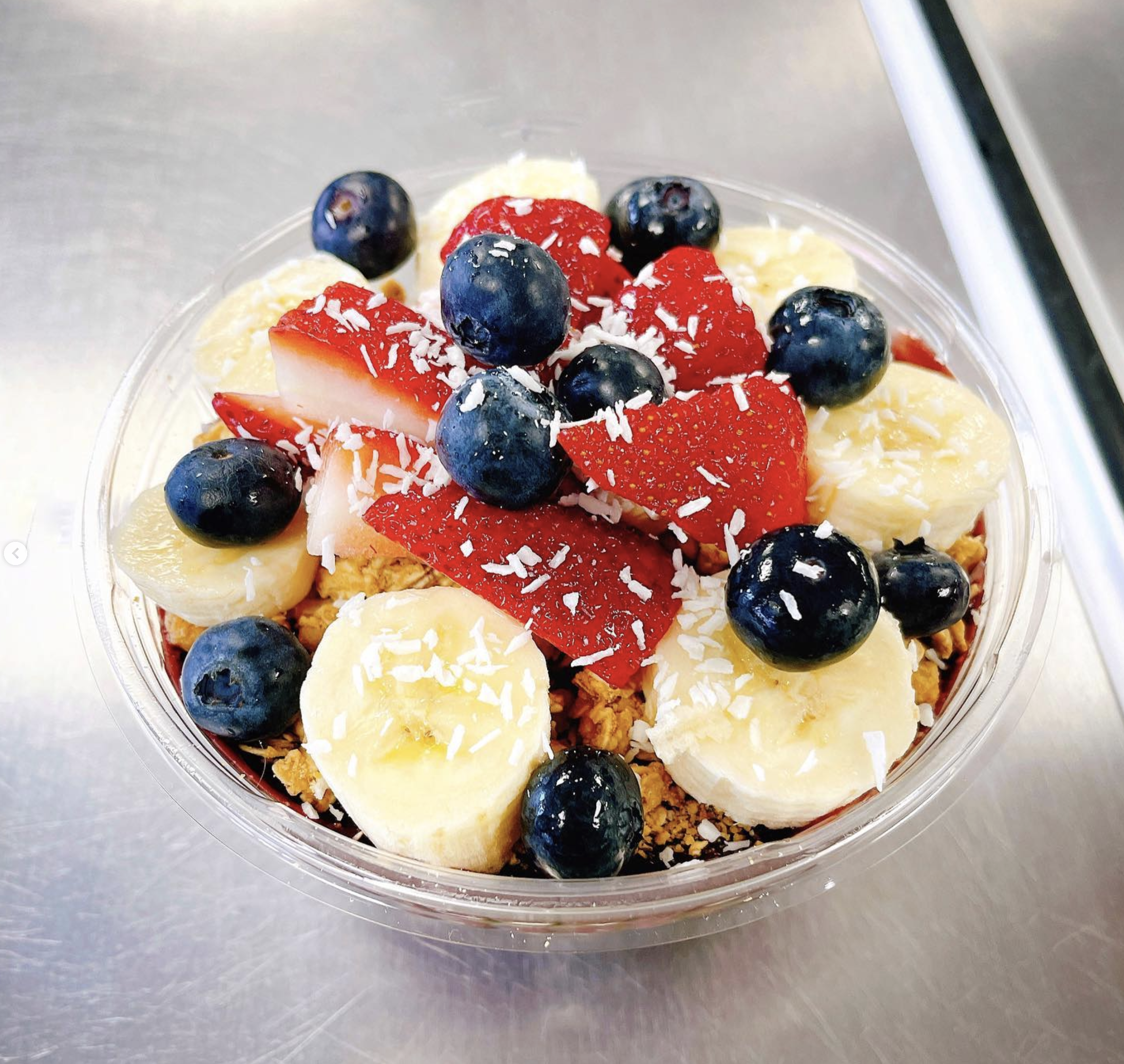 Close-up of a dessert with blueberries, sliced strawberries, banana slices, coconut flakes, and crumbly topping in a clear plastic container.