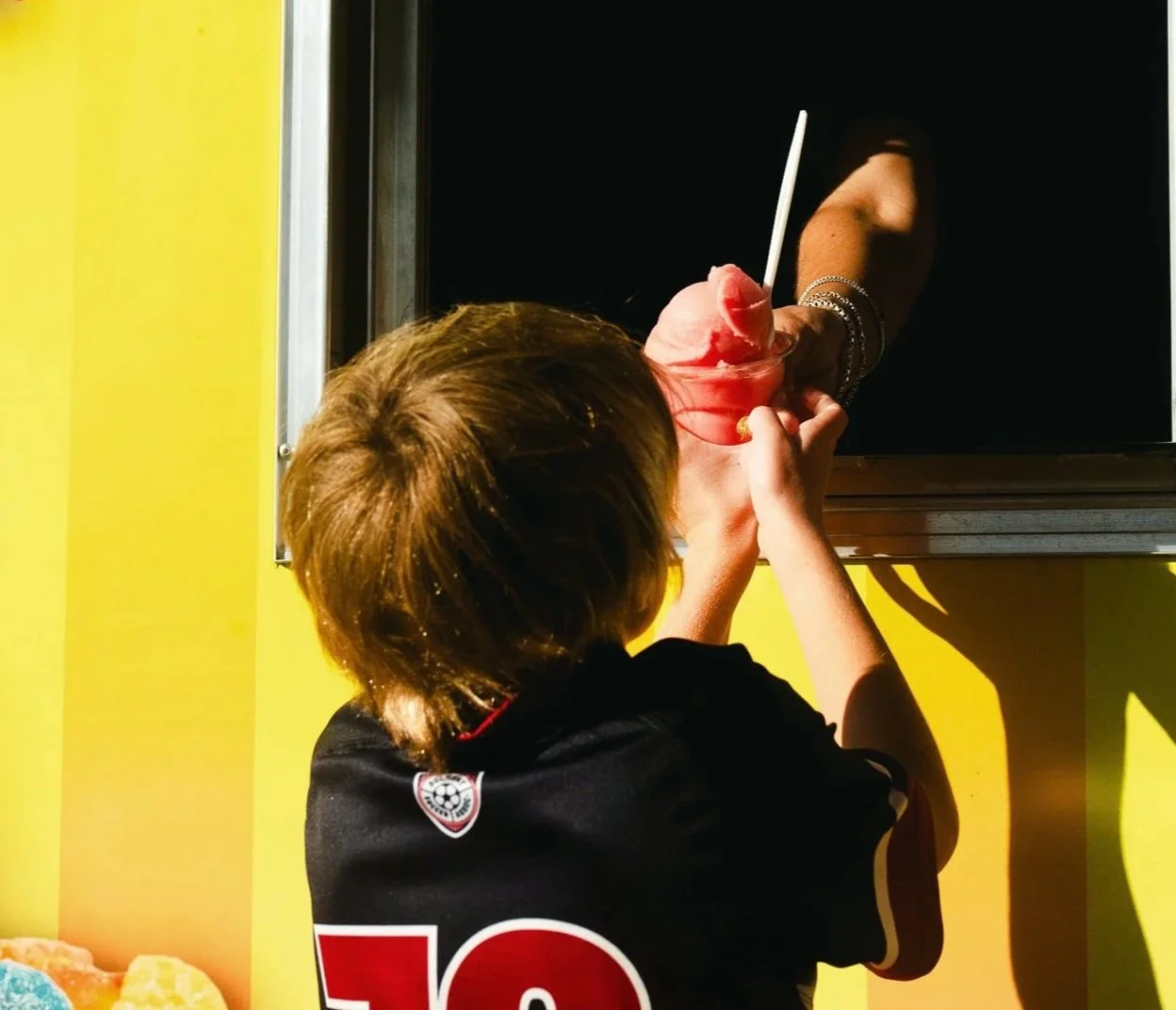 Person with light brown hair wearing a black shirt is handing a cup of pink ice cream with a white spoon to another person through a window in a yellow wall.