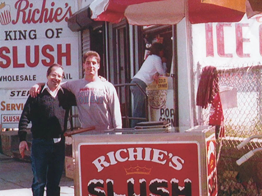 Two men standing in front of a slush stand, with a woman inside inside the stand, and a large sign that reads "Richie's" and "Slush".