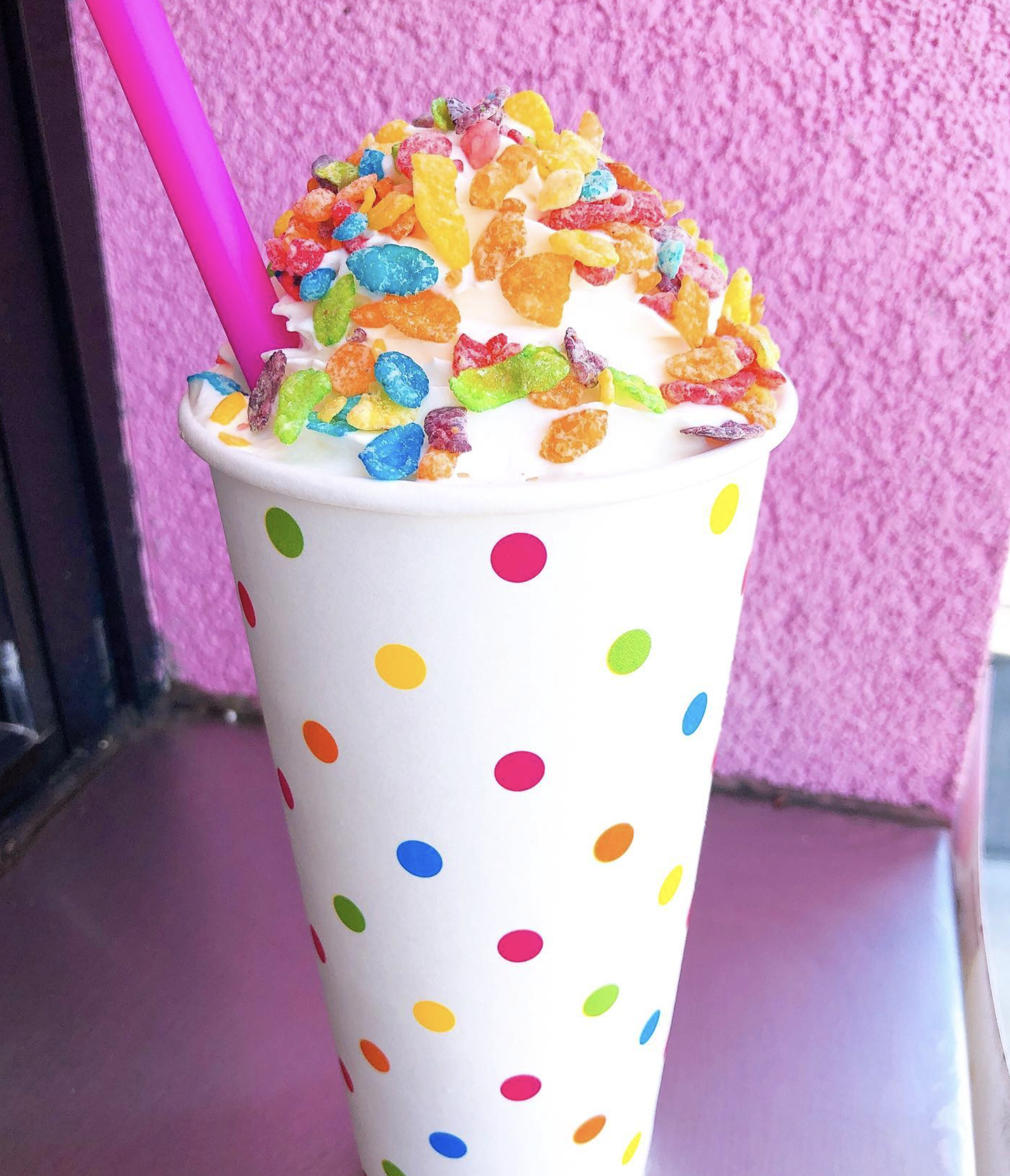 Frozen yogurt topped with colorful cereal pieces in a white cup polka-dotted with various bright colors, set against a pink textured background.