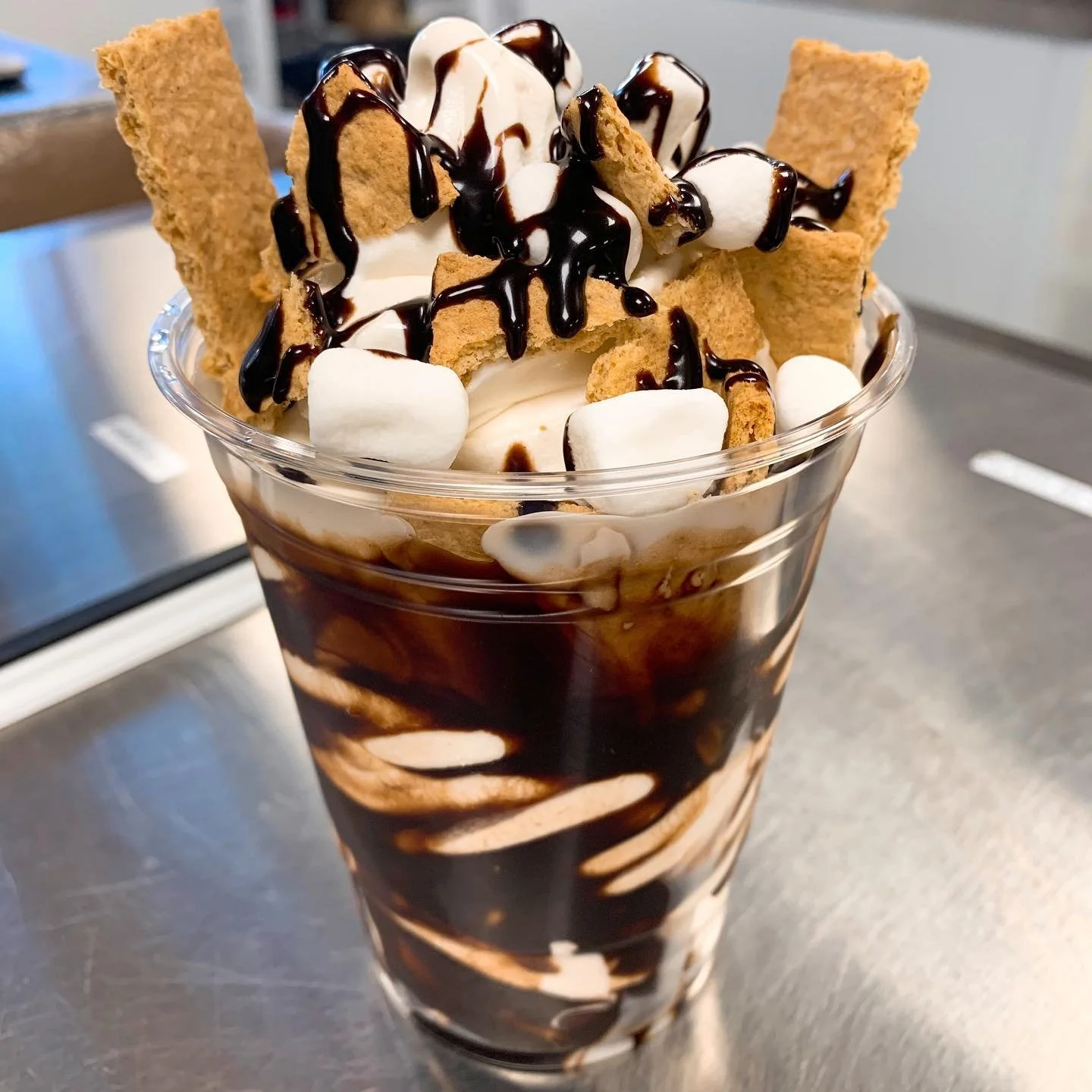 A tall glass of ice cream sunda with whipped cream, chocolate syrup, marshmallows, and graham cracker pieces on top.