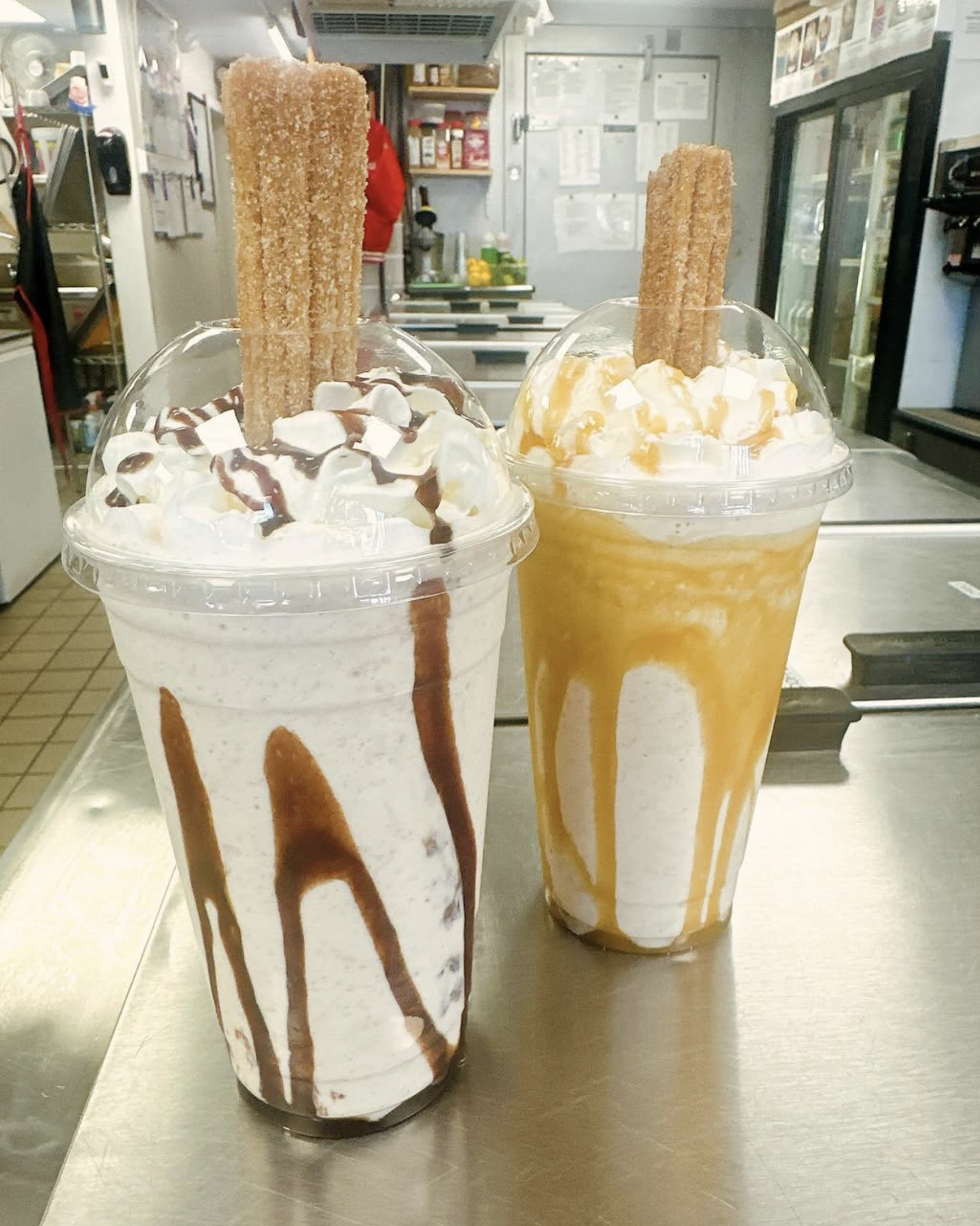 Two large milkshake drinks topped with whipped cream, caramel sauce, and chocolate drizzle, each with a churro sticking out of the whipped cream, on a metallic surface in a fast food restaurant.