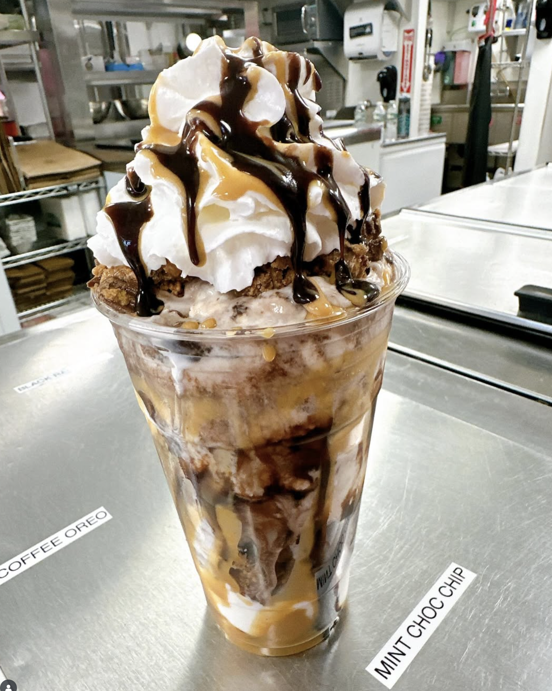 A dessert in a clear plastic cup with whipped cream, caramel, and chocolate drizzles on top, sitting on a metal counter in a kitchen.