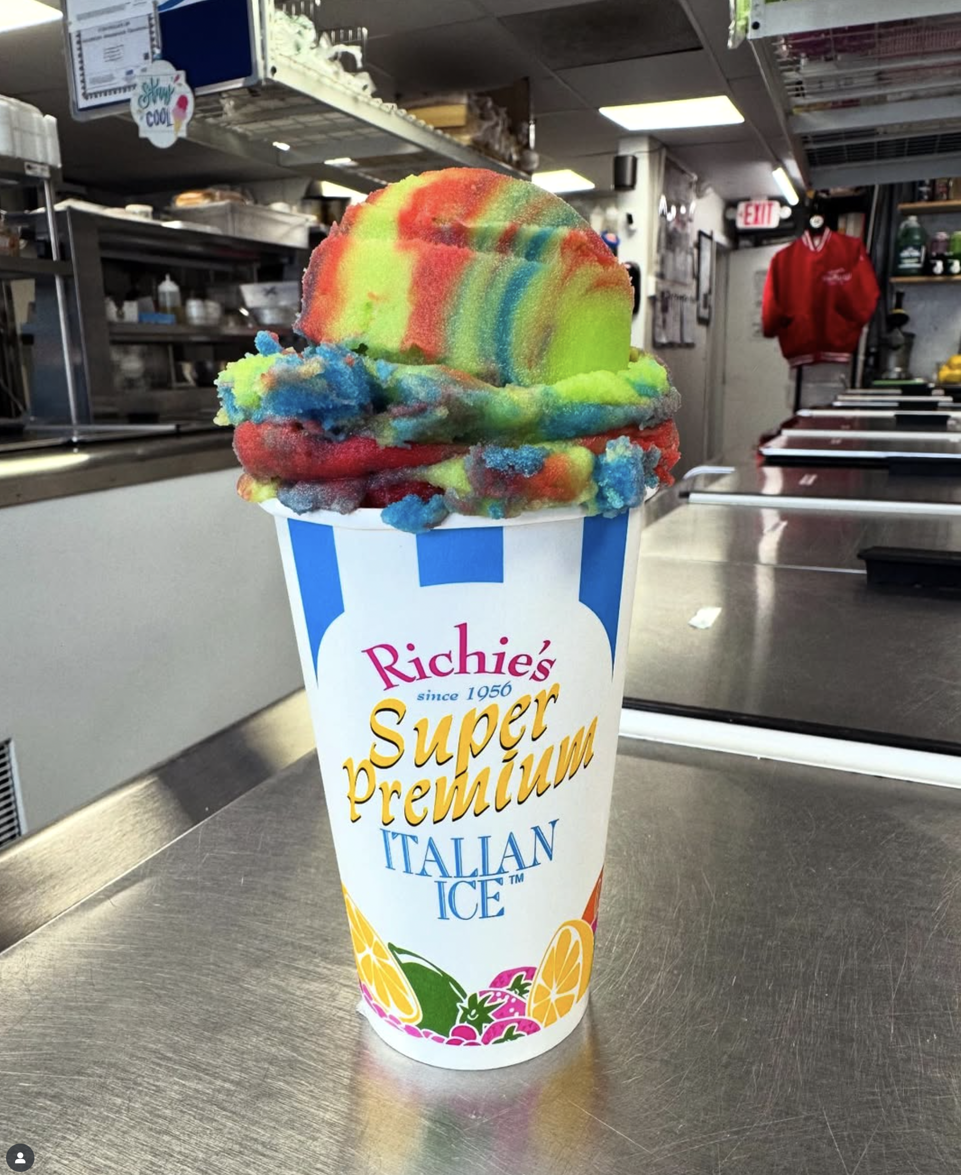 Colorful rainbow swirl ice cream in a striped cup on a metal countertop inside an ice cream shop.