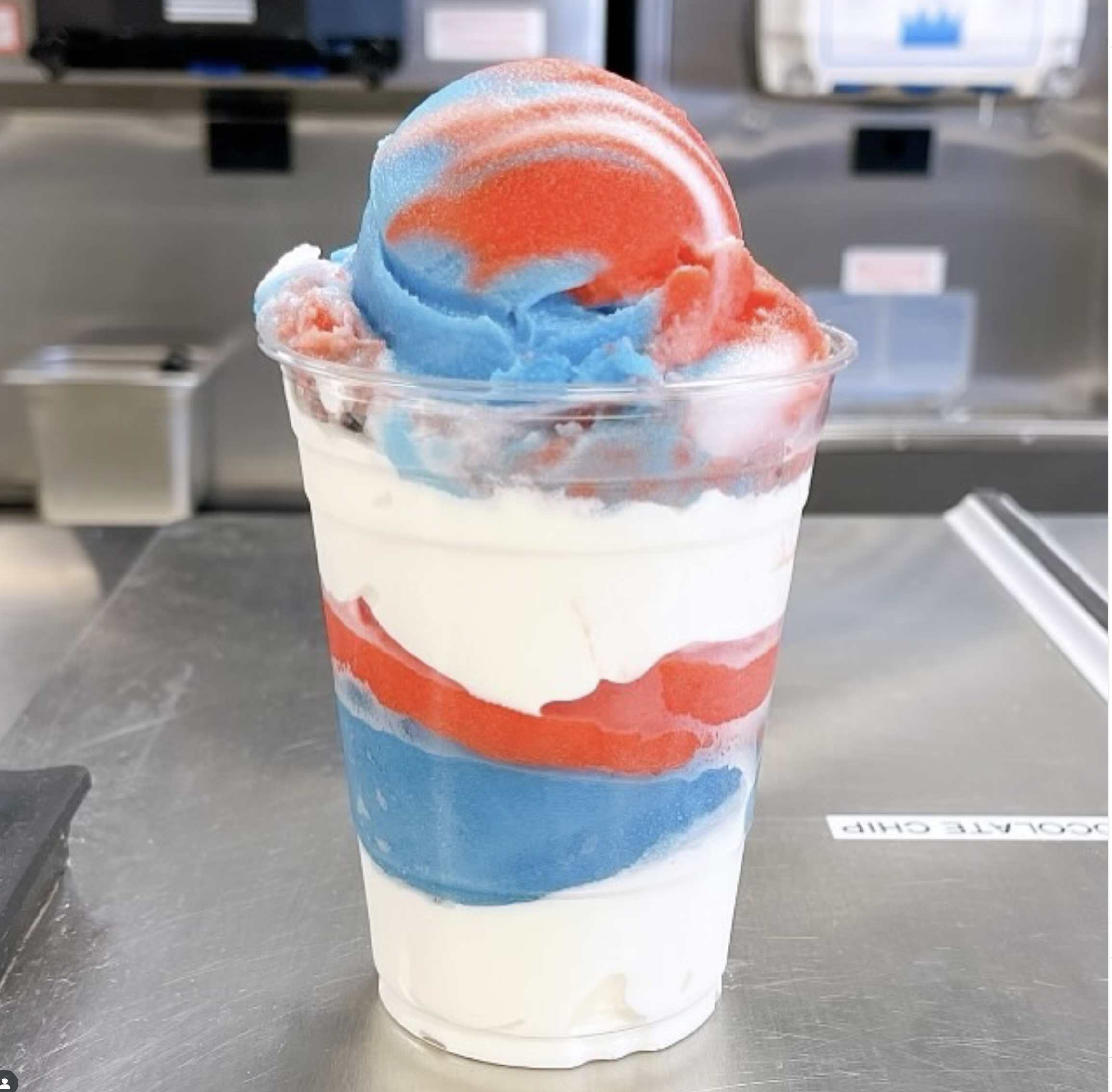 A cup of blue, red, and white shaved ice with a red orange colored sphere on top, placed on a metal counter in what appears to be an ice shop or store.