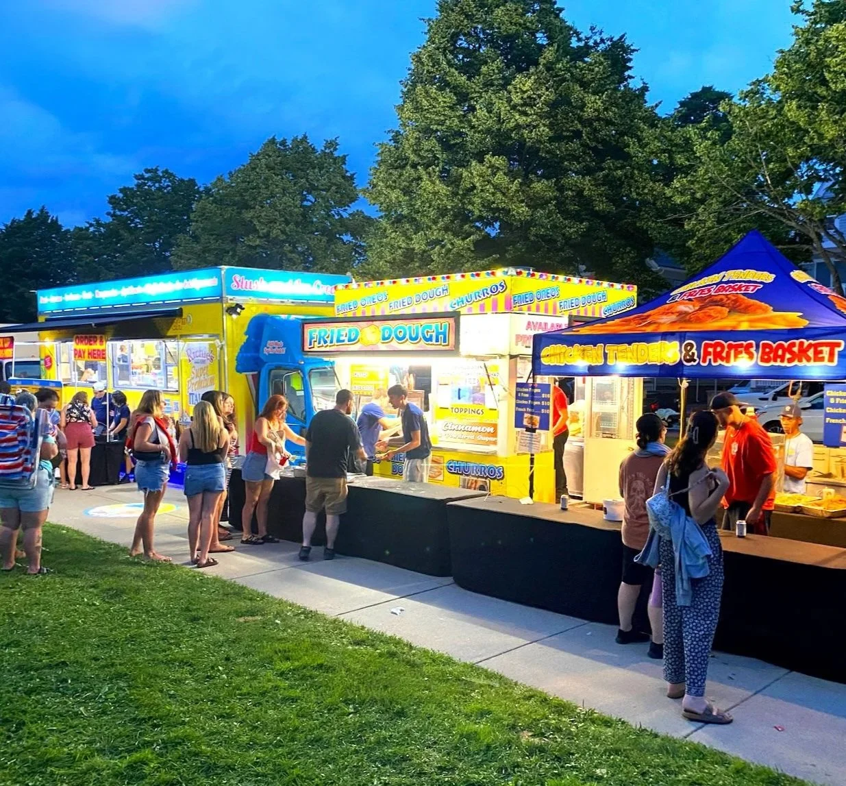 People ordering food at a fair with bright food trucks and tents, illuminated in evening light, serving fried dough, churros, chicken tenders, and fries.