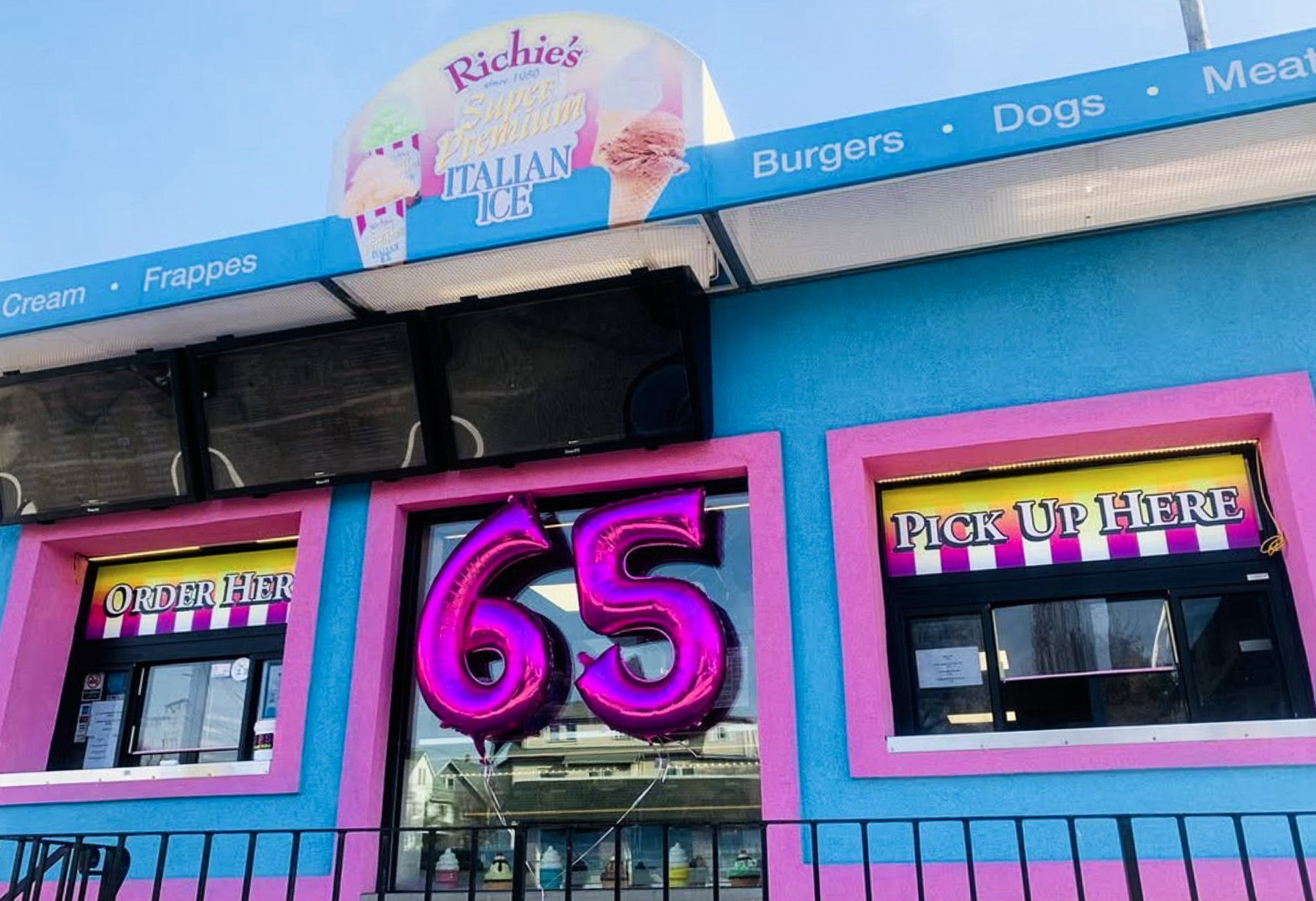 The exterior of an ice cream shop with a colorful pink, blue, and yellow facade. A neon sign reads 'Order Here' and there are balloons in the shape of the number 6. The shop offers ice cream, sundaes, and other frozen treats.